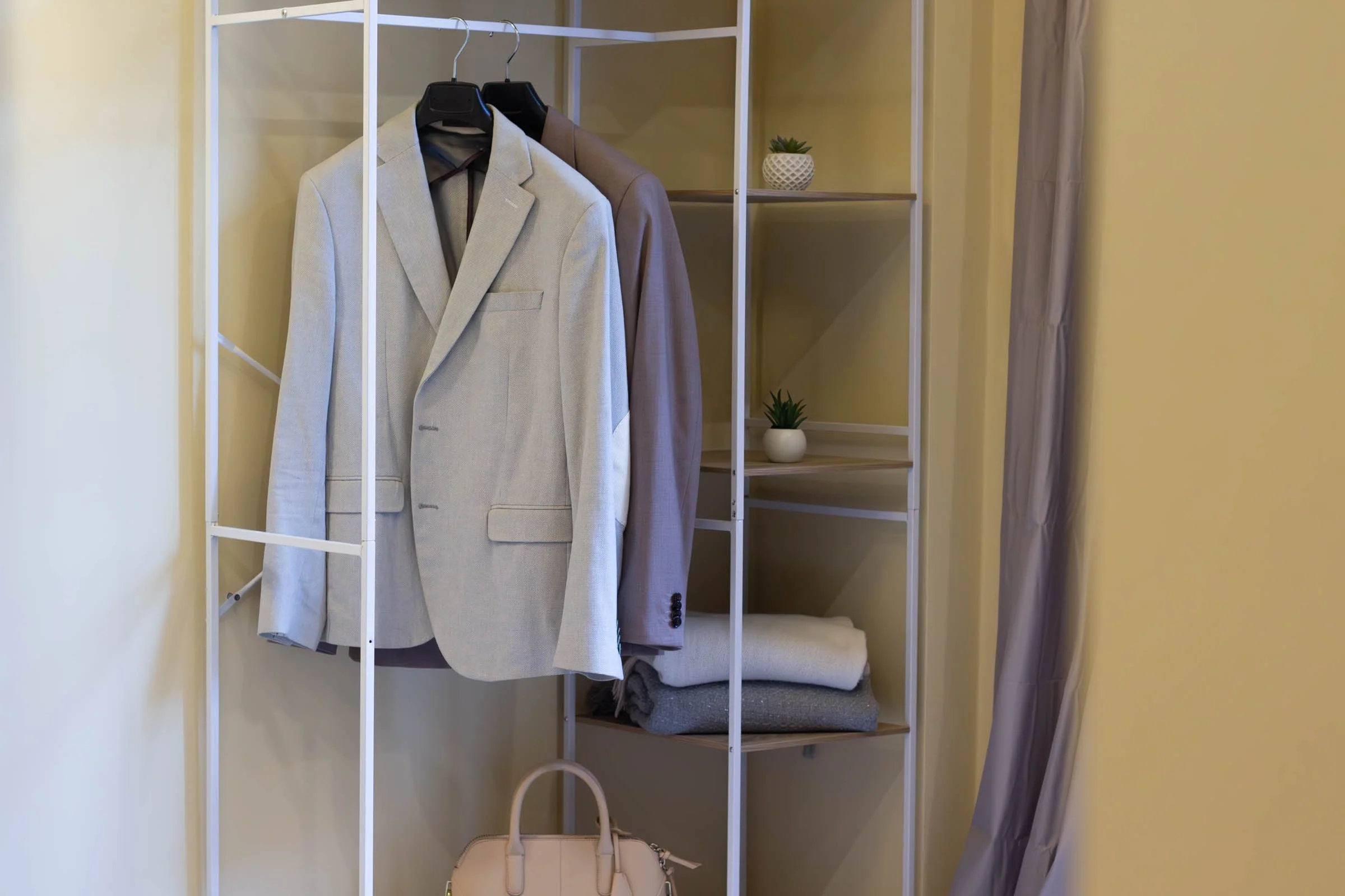 Armadio con due giacche appese, una color grigio chiaro e l'altra color beige, con piante decorative sulle mensole e una banca sotto l'armadio.