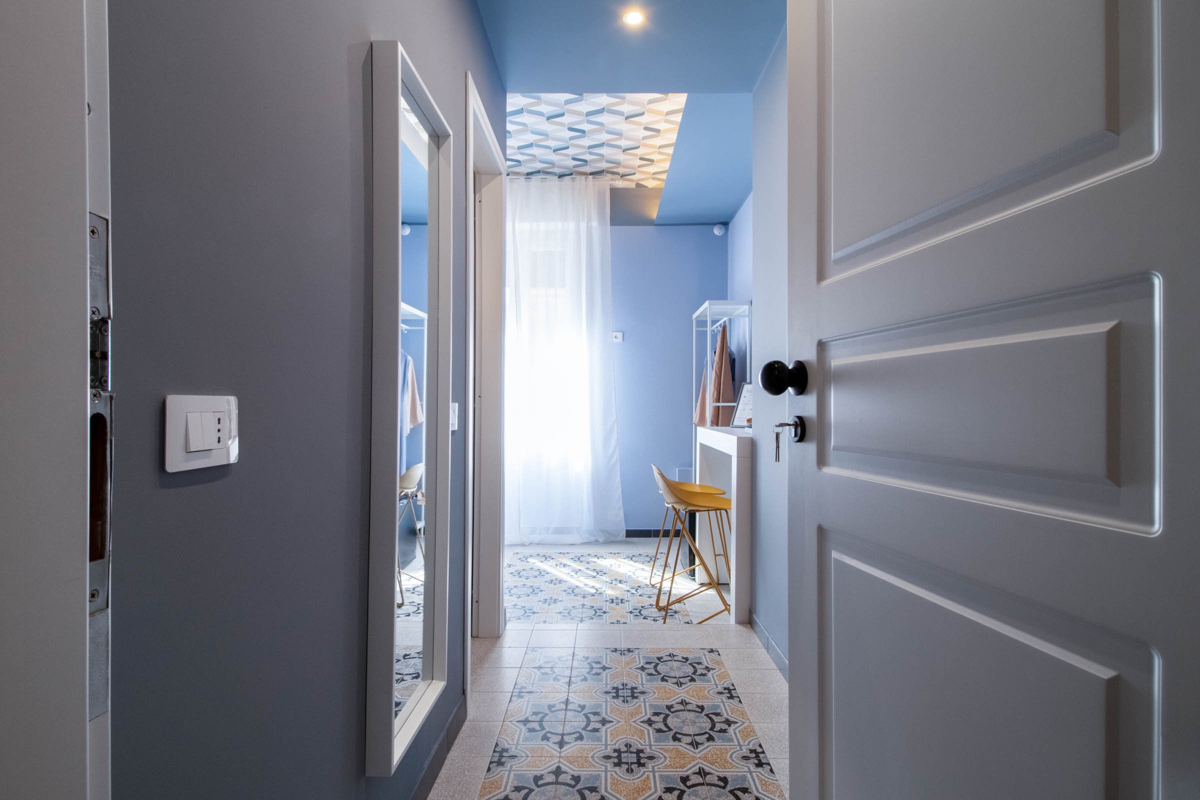 Ingresso di un appartamento con pavimento decorato, pareti azzurre, porta bianca aperta, e due sedie gialle vicino a un tavolo. In fondo, ci sono tende bianche e una stanza con pareti azzurre e un soffitto con motivo geometrico.