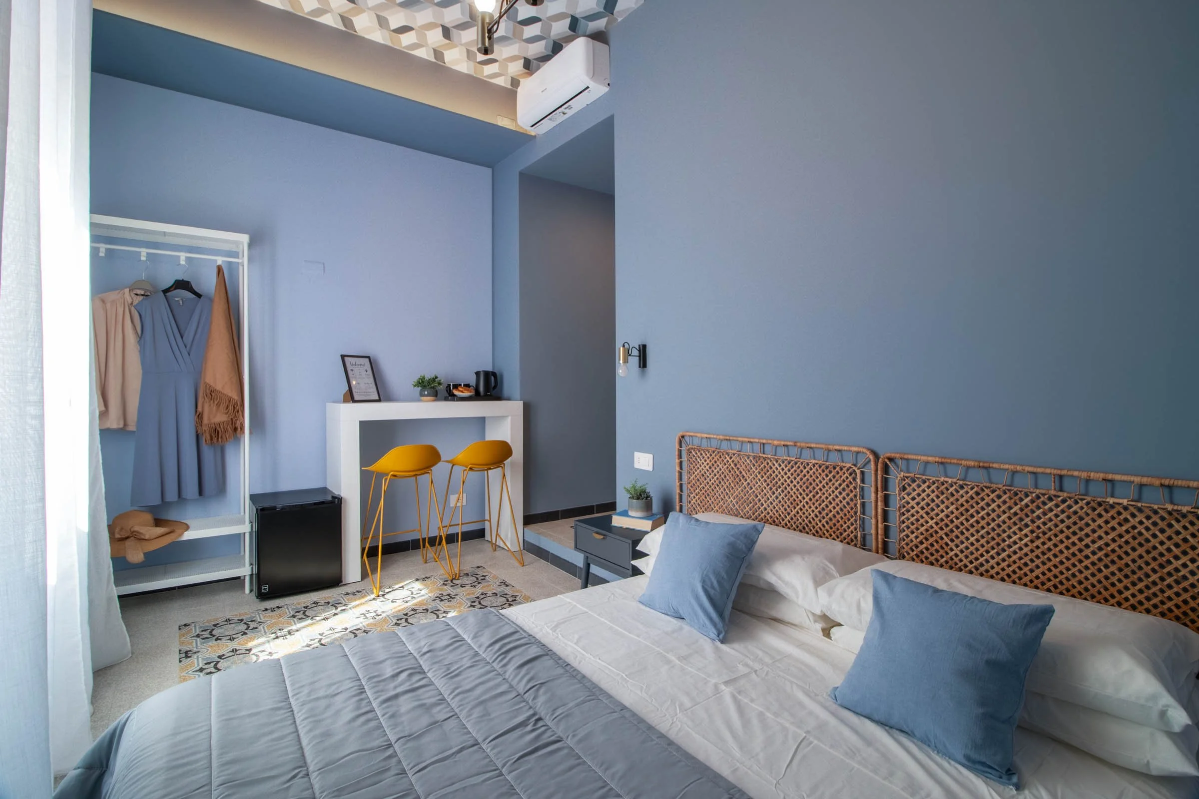 Camera da letto con due letti con cuscini blu, pareti azzurre, un armadio con vestiti e sciarpa, un mini frigorifero nero, tavolino con pianta e tazza, sedia gialla, parete con lampada e aria condizionata