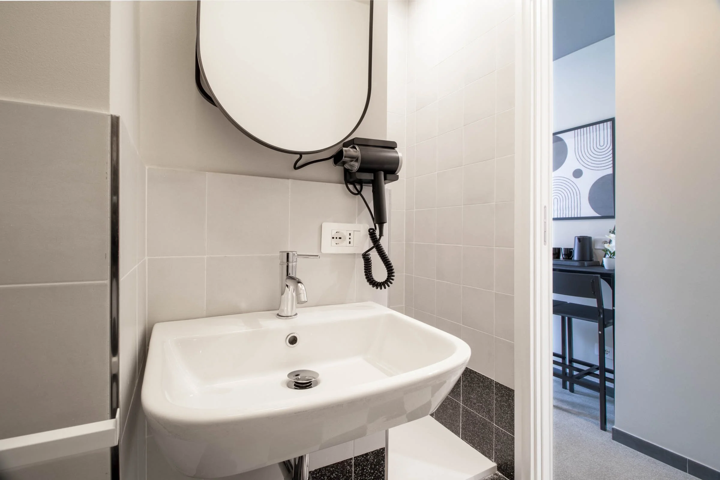 Specchio ovale sopra un lavandino bianco in un bagno moderno, con asciugacaspugliere e presa elettrica visibili, apertura che porta a una stanza con decorazioni astratte.