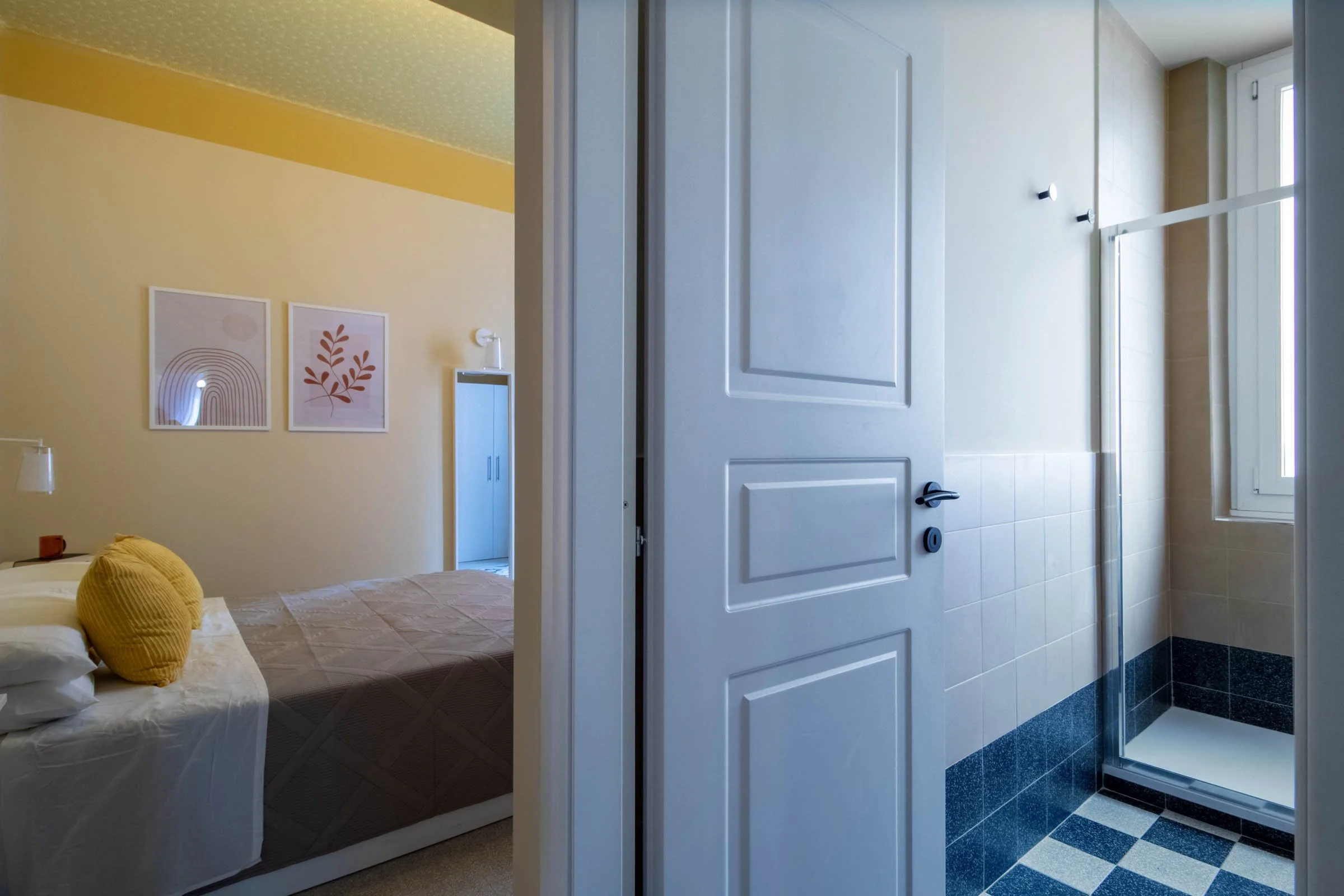 Camera da letto visibile attraverso una porta aperta, con letto con cuscini gialli, pareti gialle con due quadri e decorazioni, e una zona bagno con pareti di piastrelle e finestra.