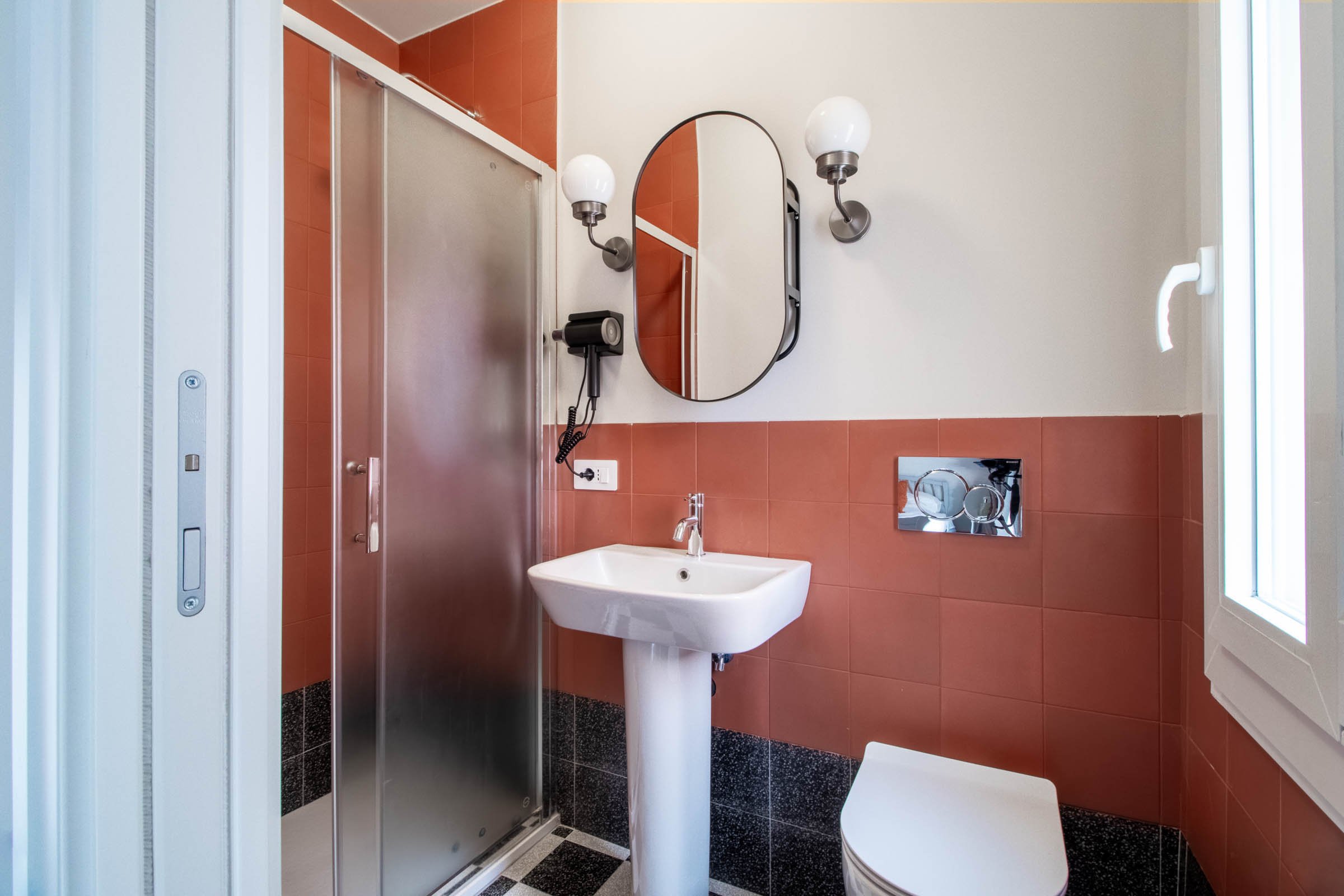 Bagno con lavabo, specchio rotondo, due luci laterali, doccia con porta in vetro, asciugamani e finestra.