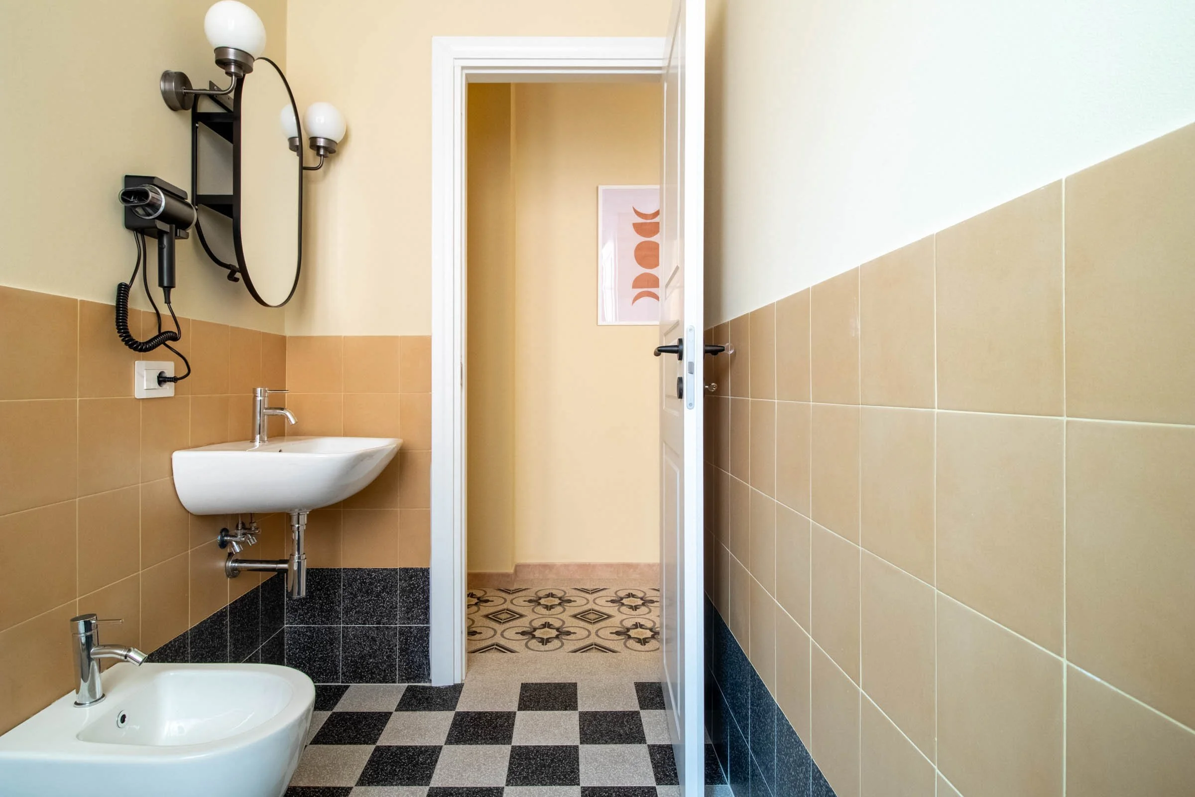 Interno di un bagno con pareti beige e piastrelle marroni e nere, con un lavandino bianco, un asciugacapelli a parete, e una porta aperta che conduce a un corridoio con pavimento decorato.