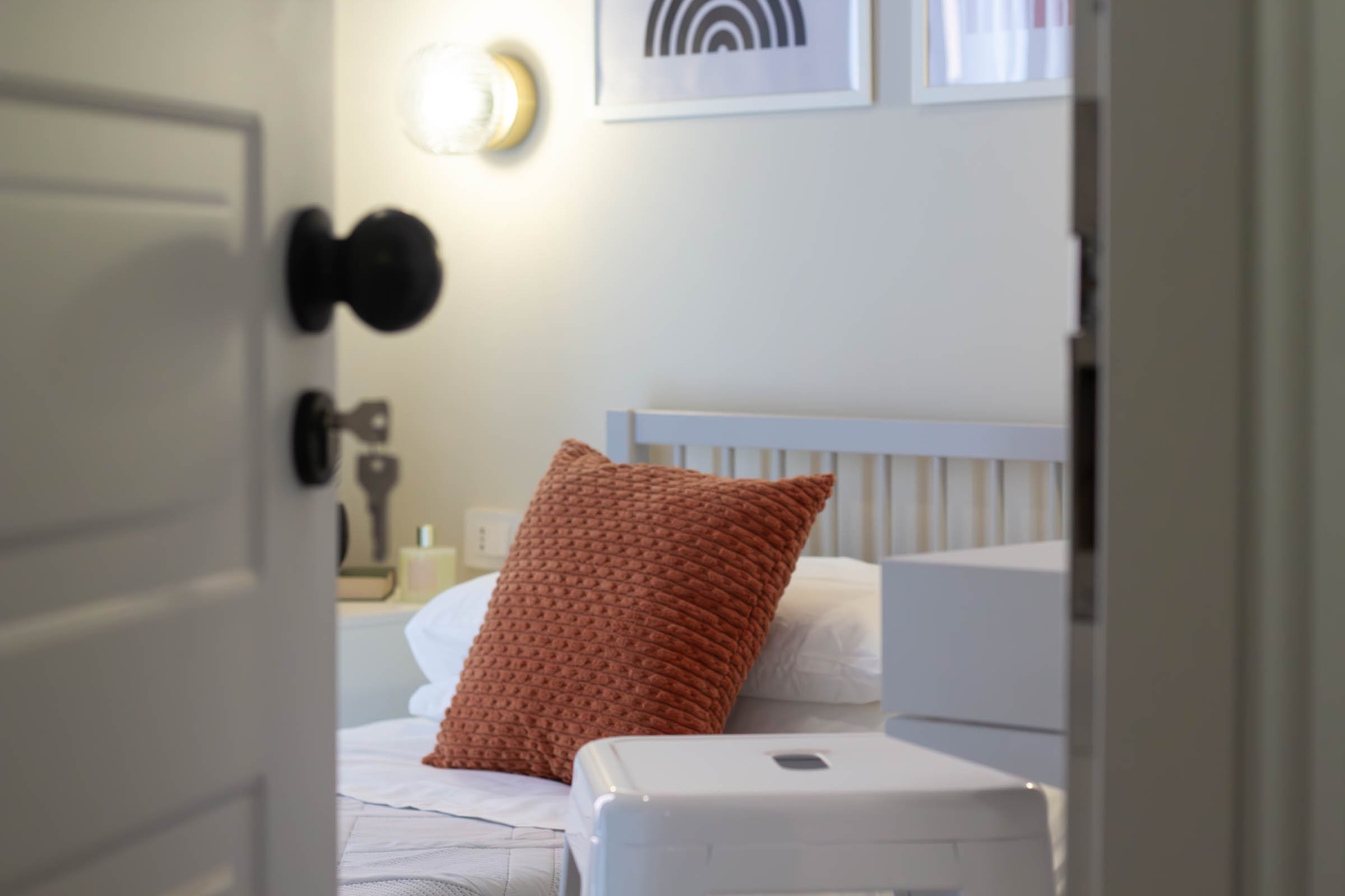 Vista di una camera da letto attraverso la soglia di una porta, con un letto con cuscini di colore arancione e una lampada sopra il comodino.