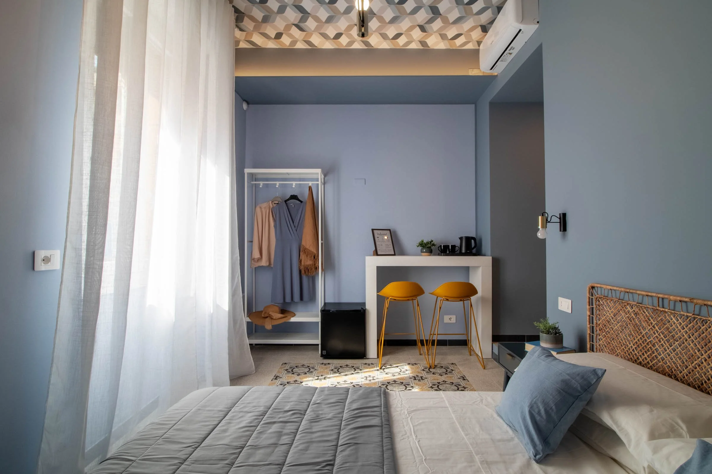 Camera da letto con pareti blu, letto con cuscini grigi, tende leggere bianche, un appendiabiti con vestiti, un tavolino con piante e tazze, sedie gialle, un'edificio con pavimento decorato e un condizionatore, stile moderno e luminoso.