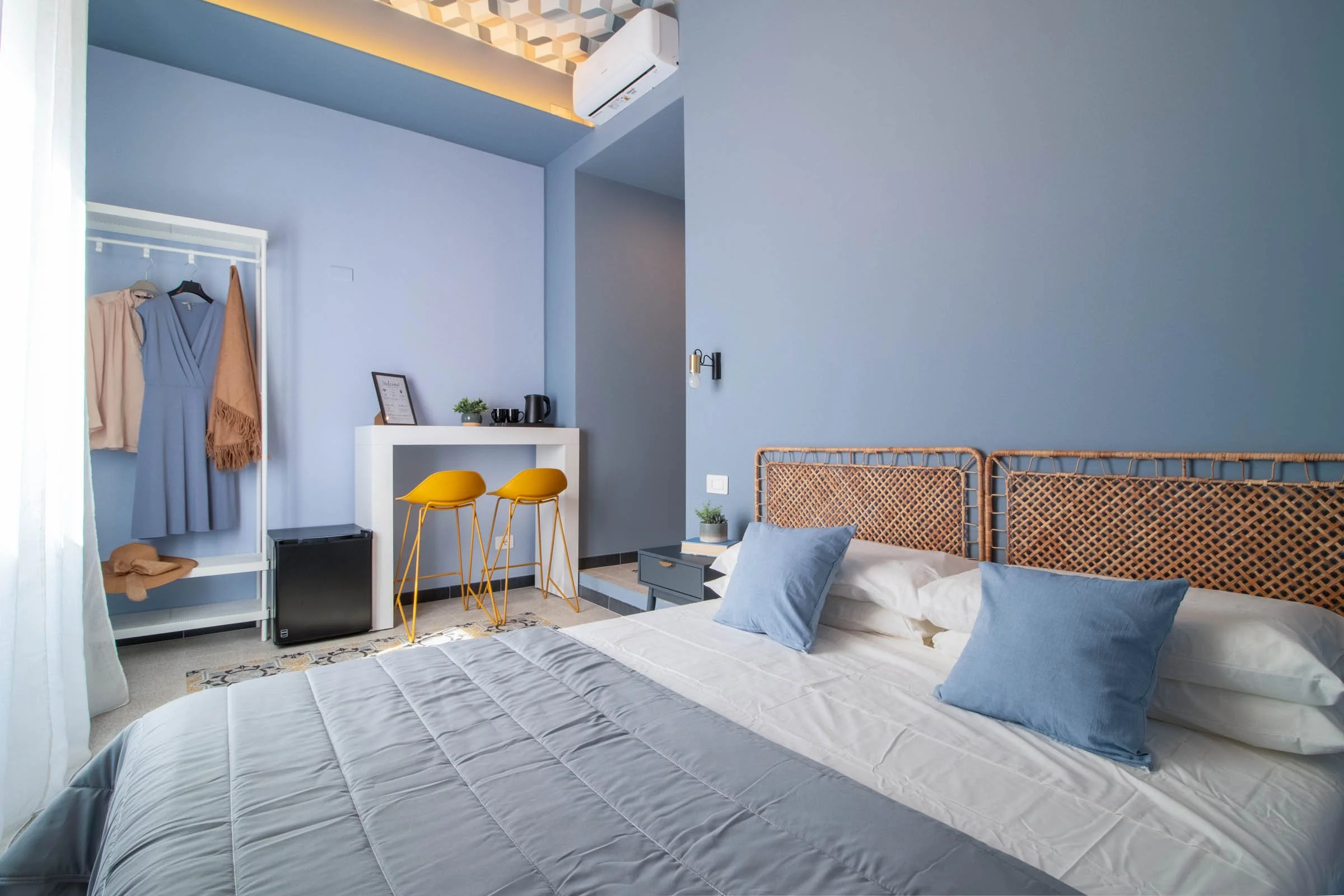 Camera da letto con pareti di colore blu chiaro, letto con cuscini azzurri e biancheria bianca. A sinistra, una zona con abiti appesi e un mini frigorifero sotto una mensola con una piante, bicchieri e una teiera. La stanza ha un'illuminazione calda 