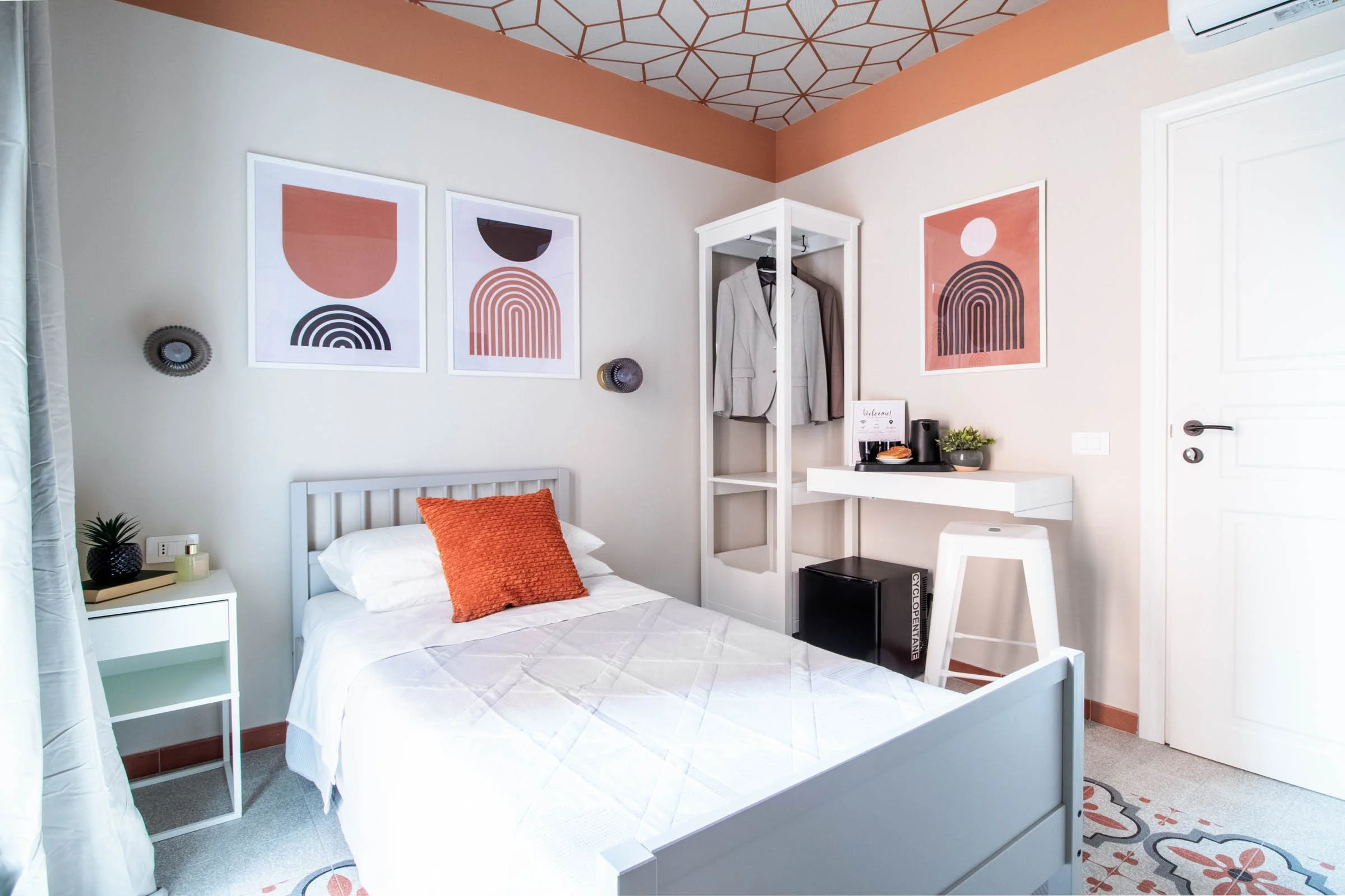 Camera da letto moderna con pareti bianche e dettagli in terracotta. Poster astratti sopra il letto, decorazioni minimaliste, comodino con pianta e libri, e un armadietto con abiti appesi. Pavimento con piastrelle colorate. Tende leggere e aria condi
