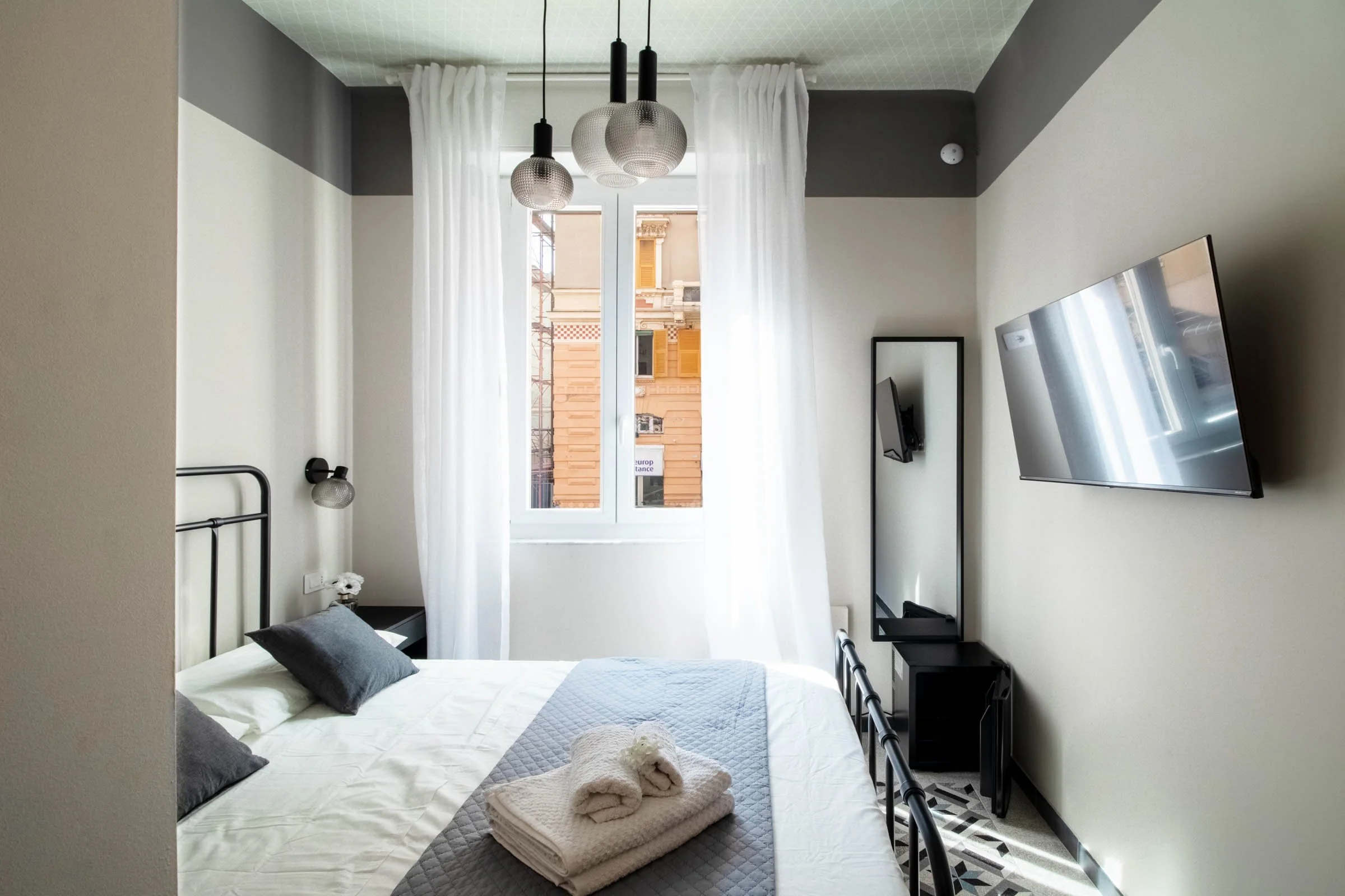 Camera da letto moderna con letto, cuscini grigi, soffitto con luci pendenti a sfera, finestra con tende bianche, televisore a parete, e pavimento con piastrelle geometriche.