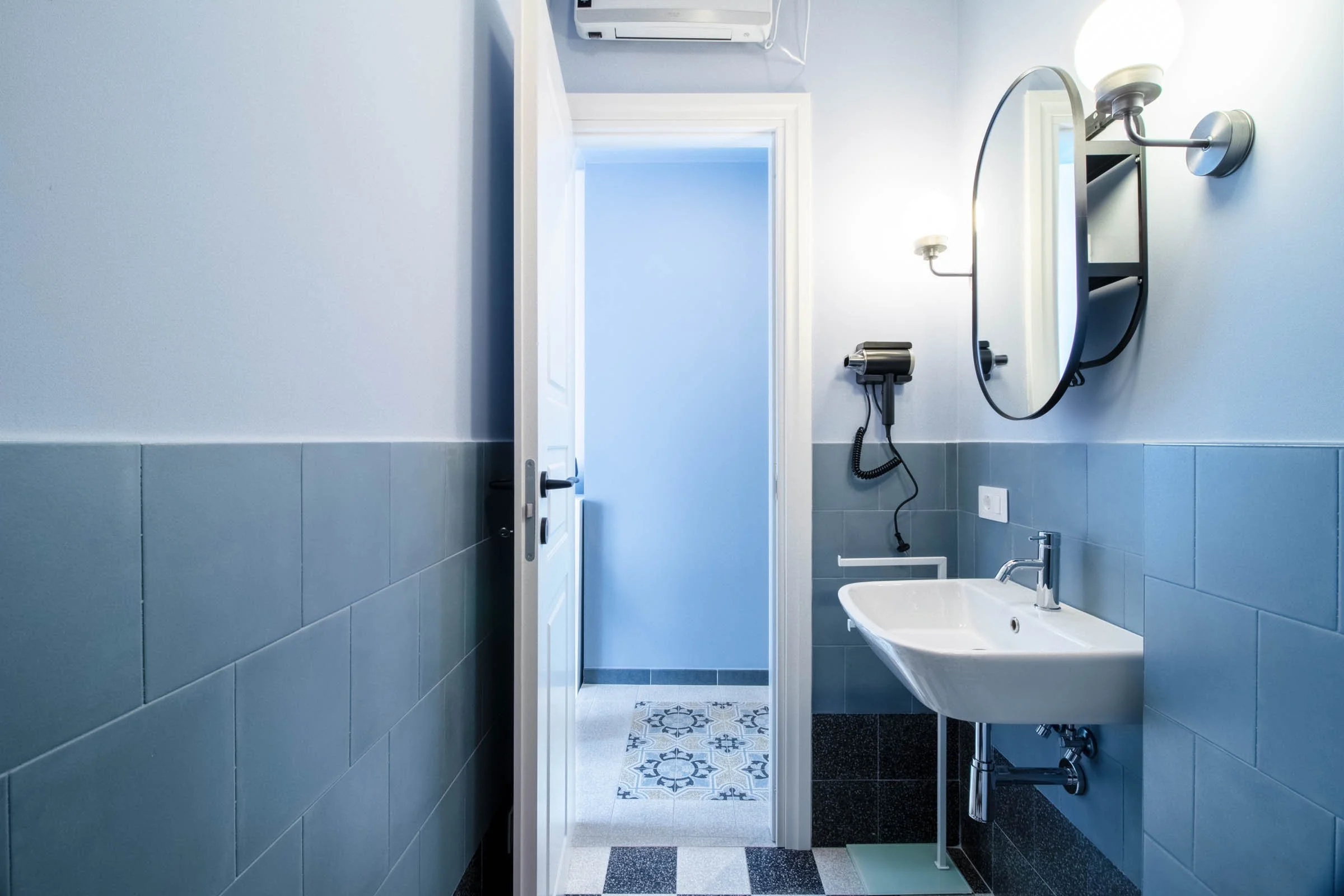 Bagno con pareti azzurre, lavabo bianco, specchio rotondo, lampade da parete, asciugacapelli, aria condizionata, porta aperta verso un'altra stanza, pavimento con piastrelle decorative.