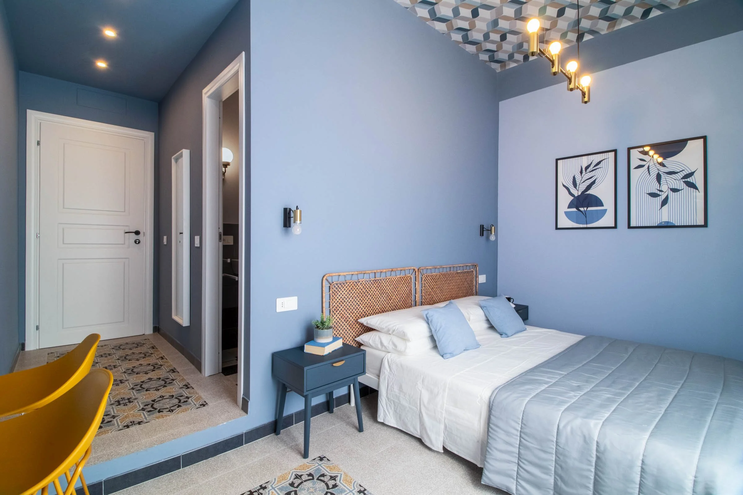 Camera da letto con pareti azzurre, letto con lenzuola bianche e cuscini azzurri, quadri astratti alle pareti, lampade a muro, tavolino blu con libri e pianta, arredamento moderno.