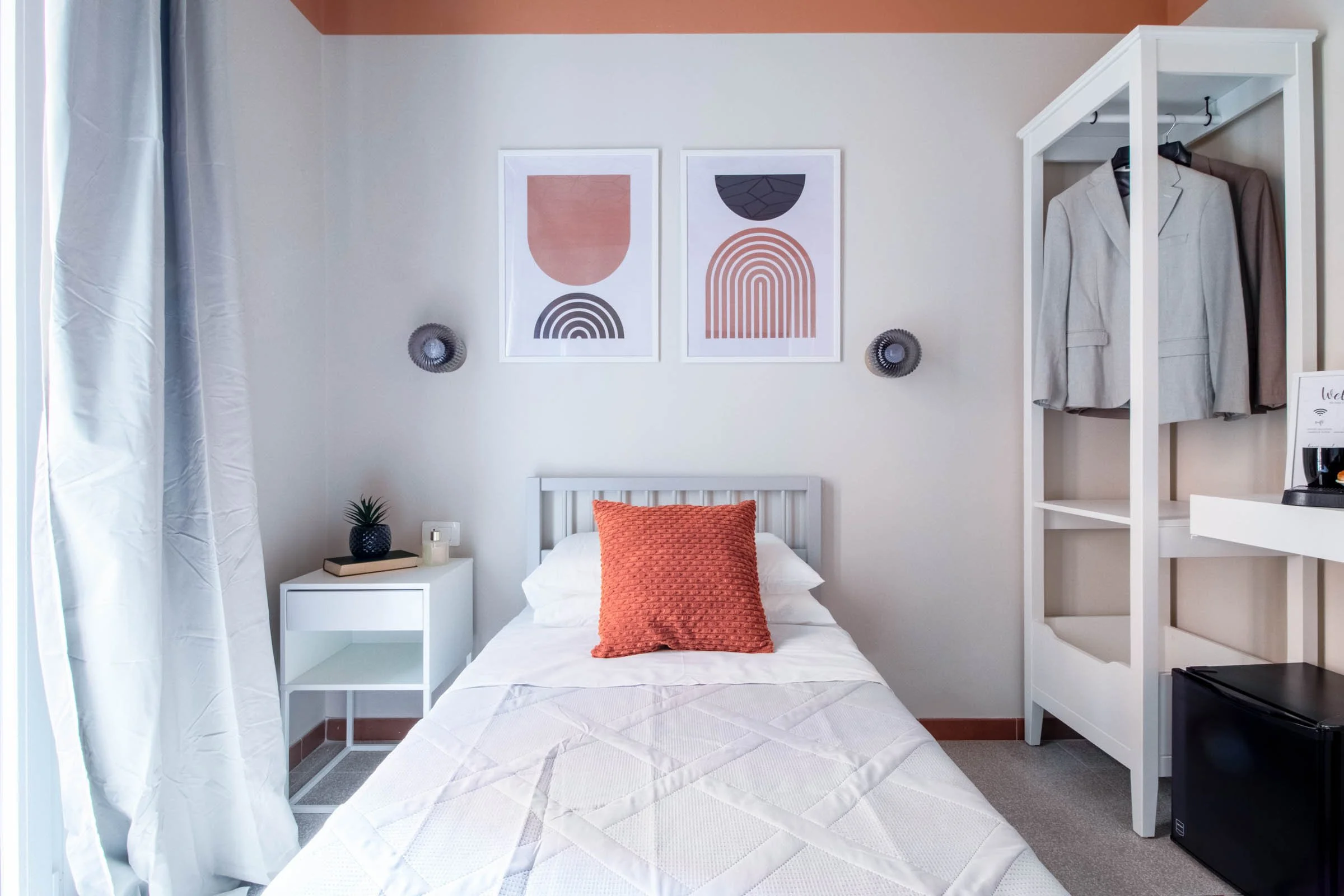 Camera da letto con pareti bianche e dettagli color pesca. Letto con copriletto bianco e cuscino arancione. Sul muro, due quadri astratti con motivi geometrici e colori neutri. Accanto al letto, una piccola cassettiera con decorazioni e una pianta in