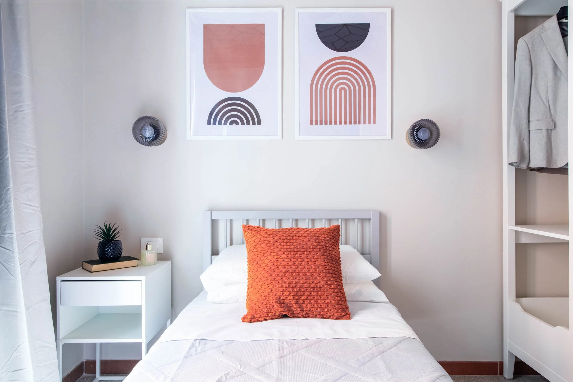 Camera da letto con pareti bianche, decorata con due quadri astratti con motivi geometrici e colori rosa, nero e beige. Sulla parete sopra il letto ci sono due faretti a forma di ventaglio nero. Sul comodino bianco a sinistra sono presenti una pianta