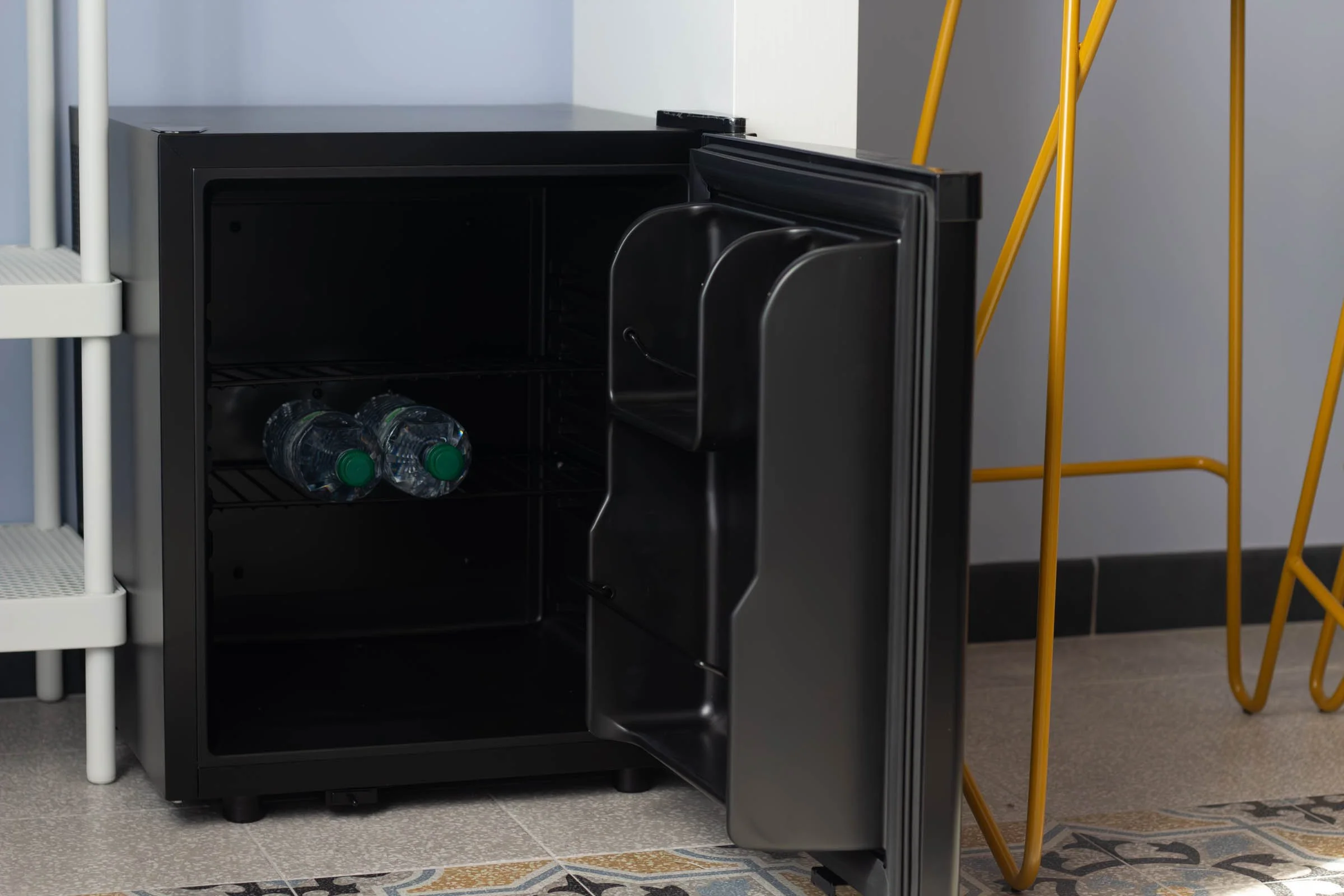 Mini frigo nero aperto con alcune bottiglie d'acqua all'interno, situato in un ambiente con pavimenti di piastrelle e mobili gialli