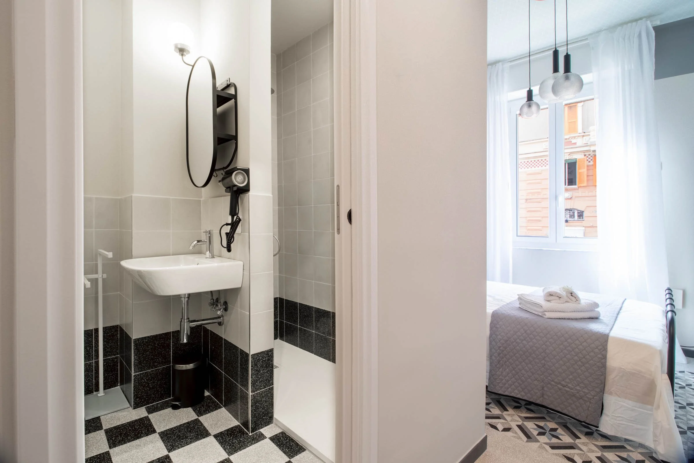 Interno di un appartamento con bagno e camera da letto; il bagno ha piastrelle bianche e nere, lavabo e specchio, la camera mostra un letto con asciugamani e vista su un edificio con balcone.