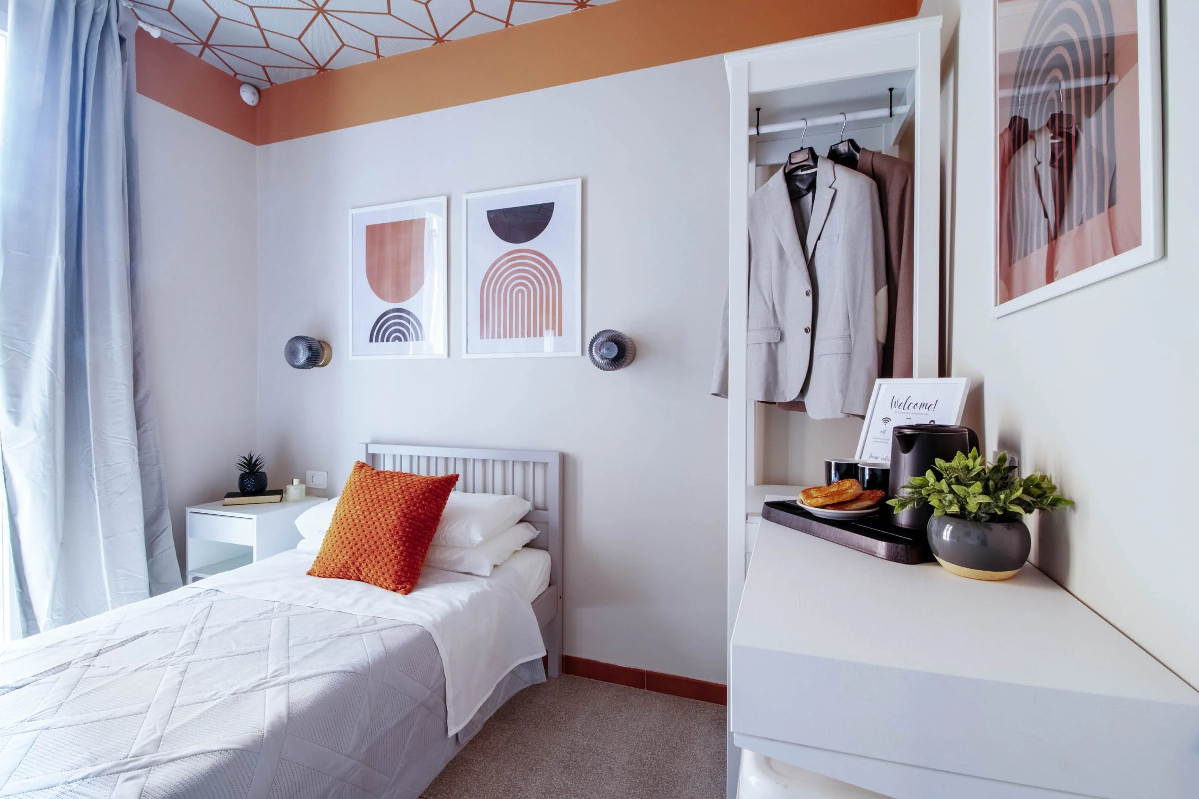 Camera da letto moderna con letto bianco, cuscino arancione e quadri astratti sulla parete, scaffale con giacca beige e altre decorazioni, decorazioni floreali e una zona colazione con toast e macchina del caffè.