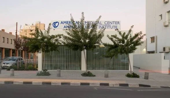 A street view showing the American Medical Center and American Medical Institute building with a sign on the roof, three trees planted along the sidewalk, and a parked car to the side.