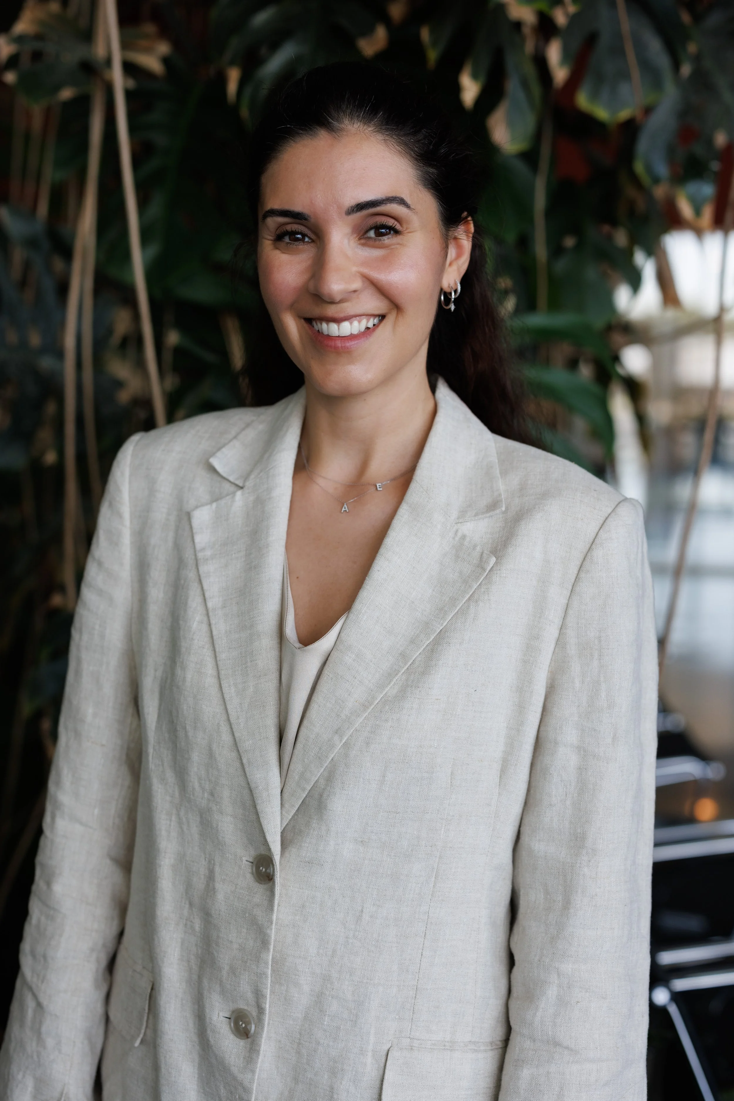 Dr Thoukididou wearing a beige blazer and jewelry, standing in front of green plants.