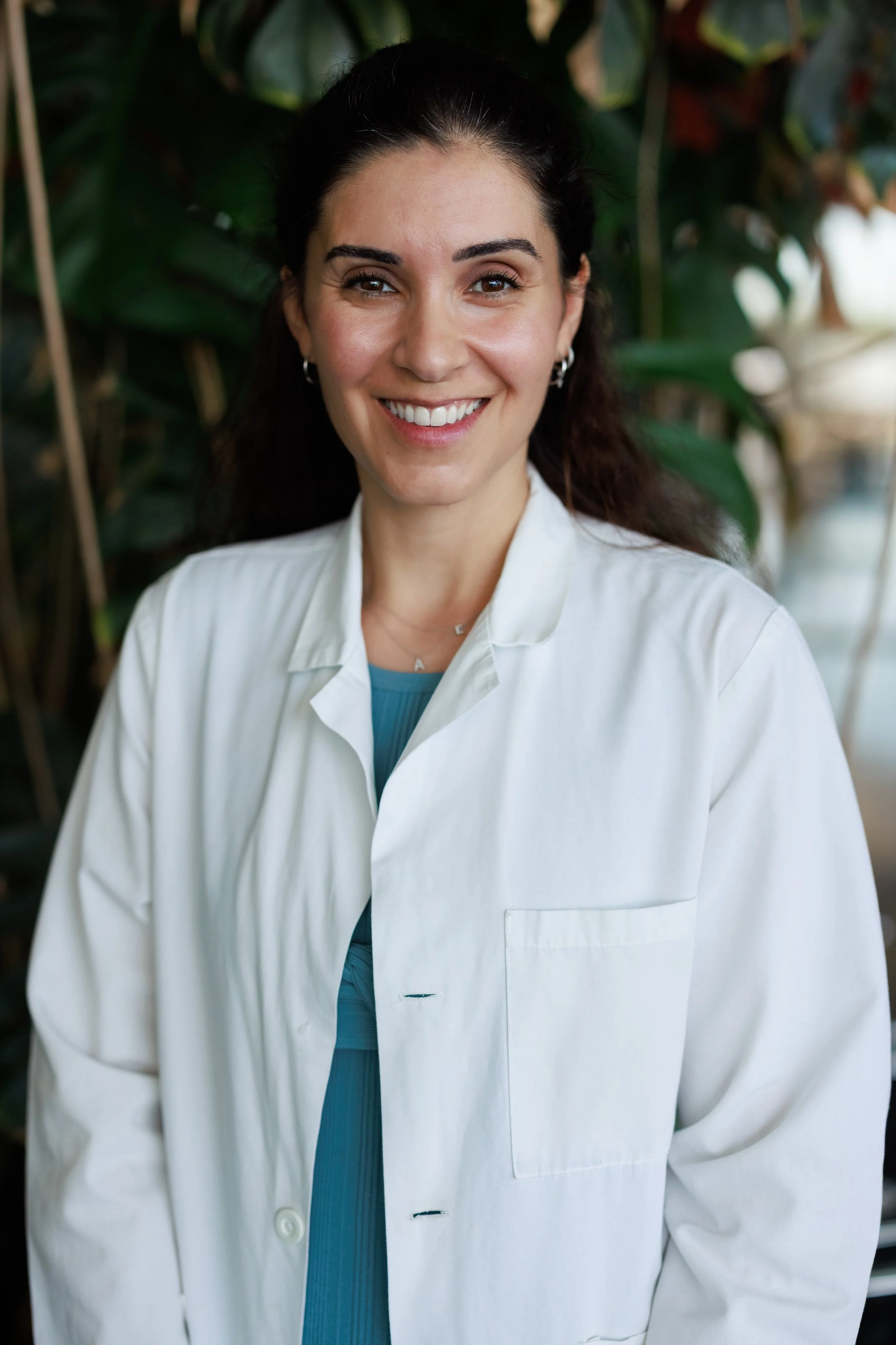 Dr Thoukididou smiling in a white lab coat, standing in front of green foliage.