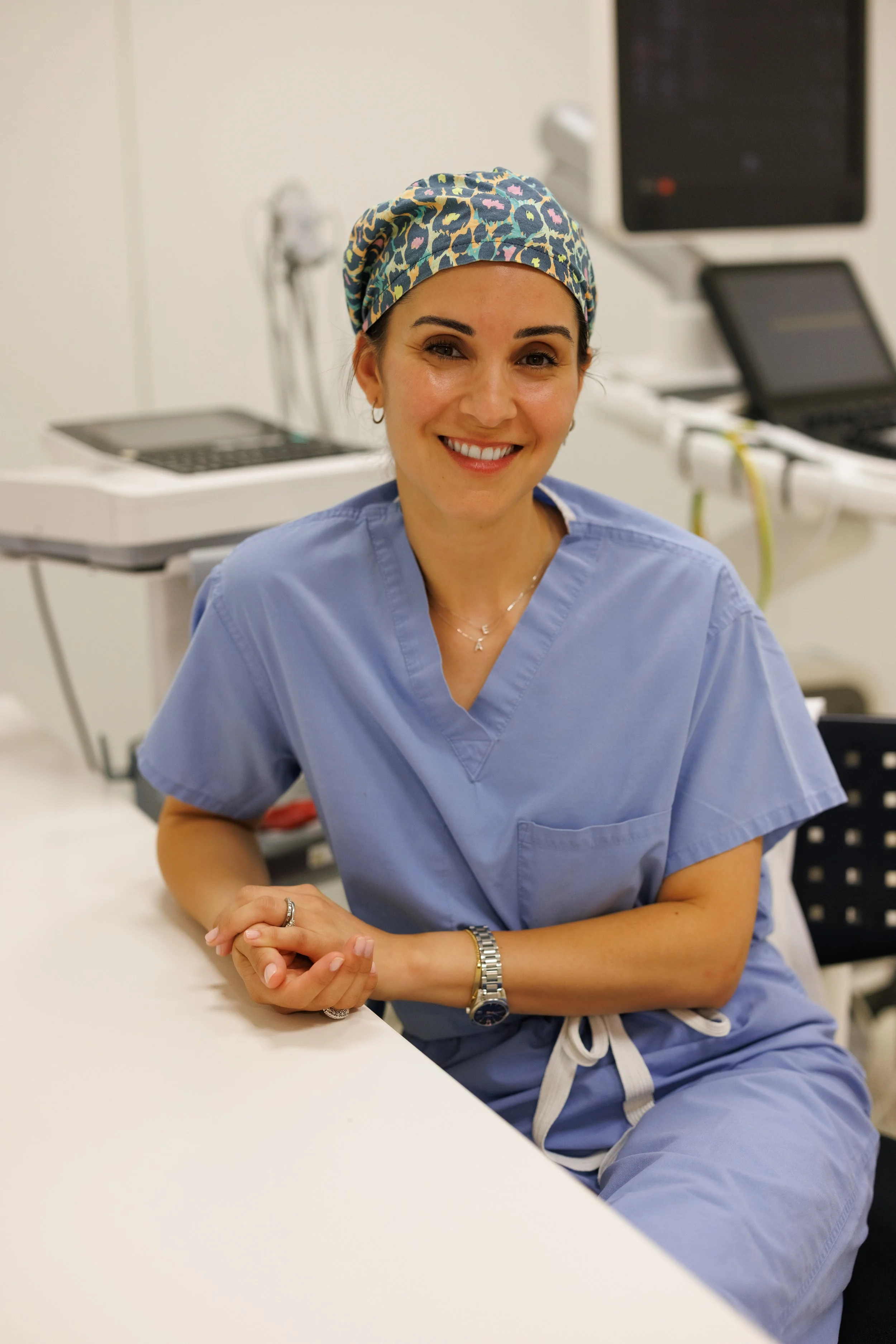 A woman in medical scrubs and a colorful headscarf, sitting at a white table in a clinical setting, smiling at the camera.
