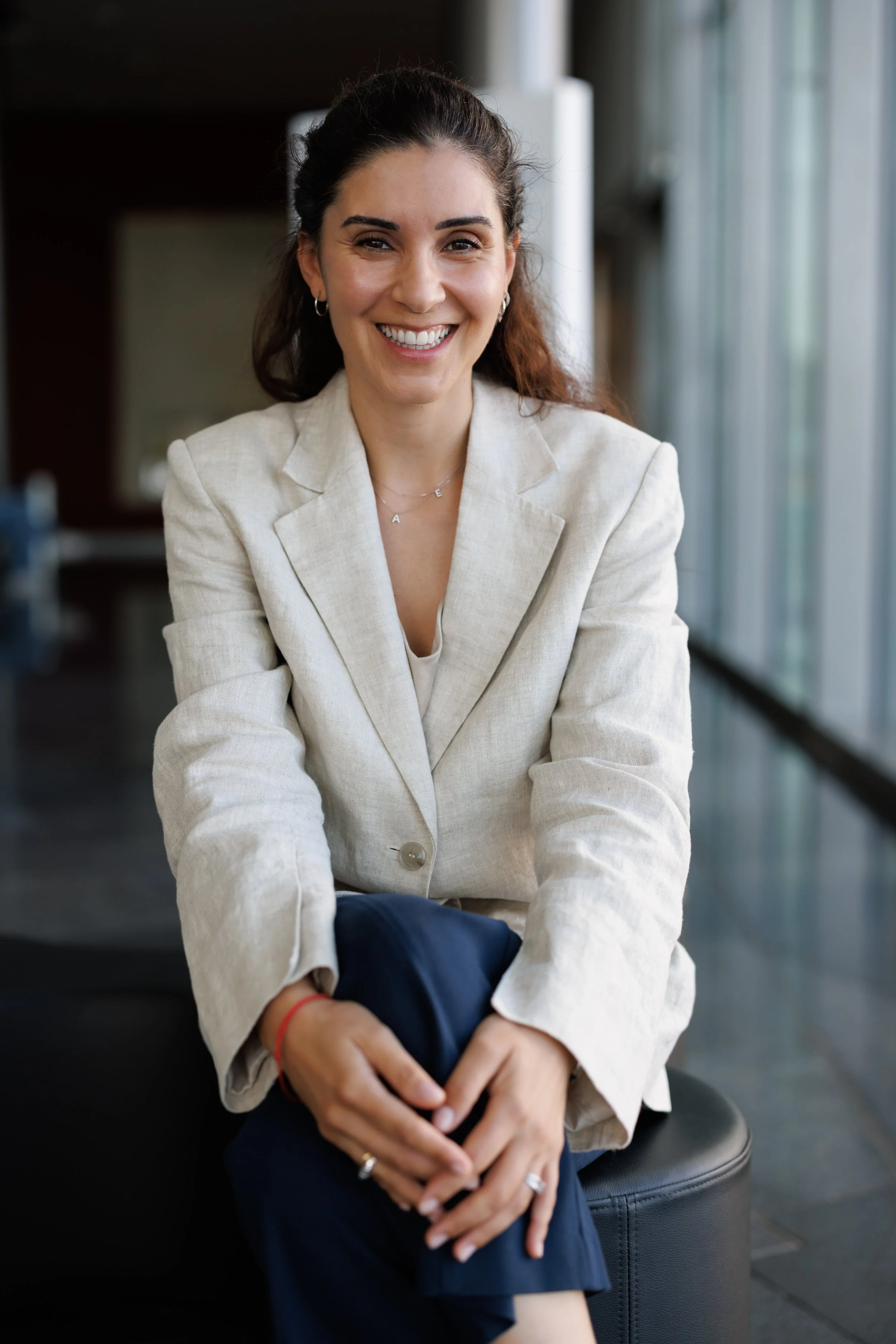 Dr Thoukdiidou with dark hair tied back, wearing a light-colored blazer and a necklace, sitting indoors with her hands resting on her lap.