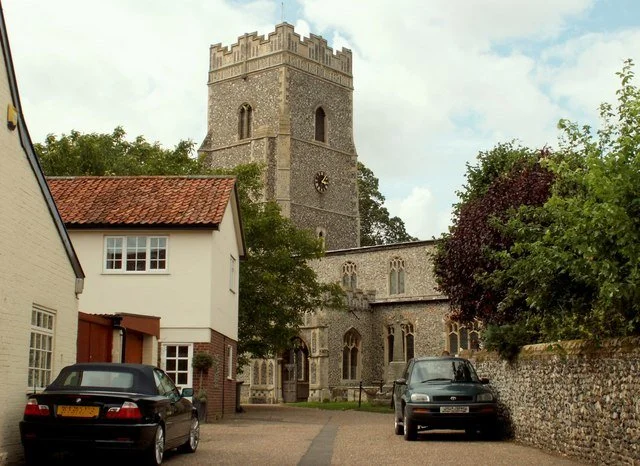 A stone church with a tall clock tower, surrounded by trees and parked cars in a small courtyard.