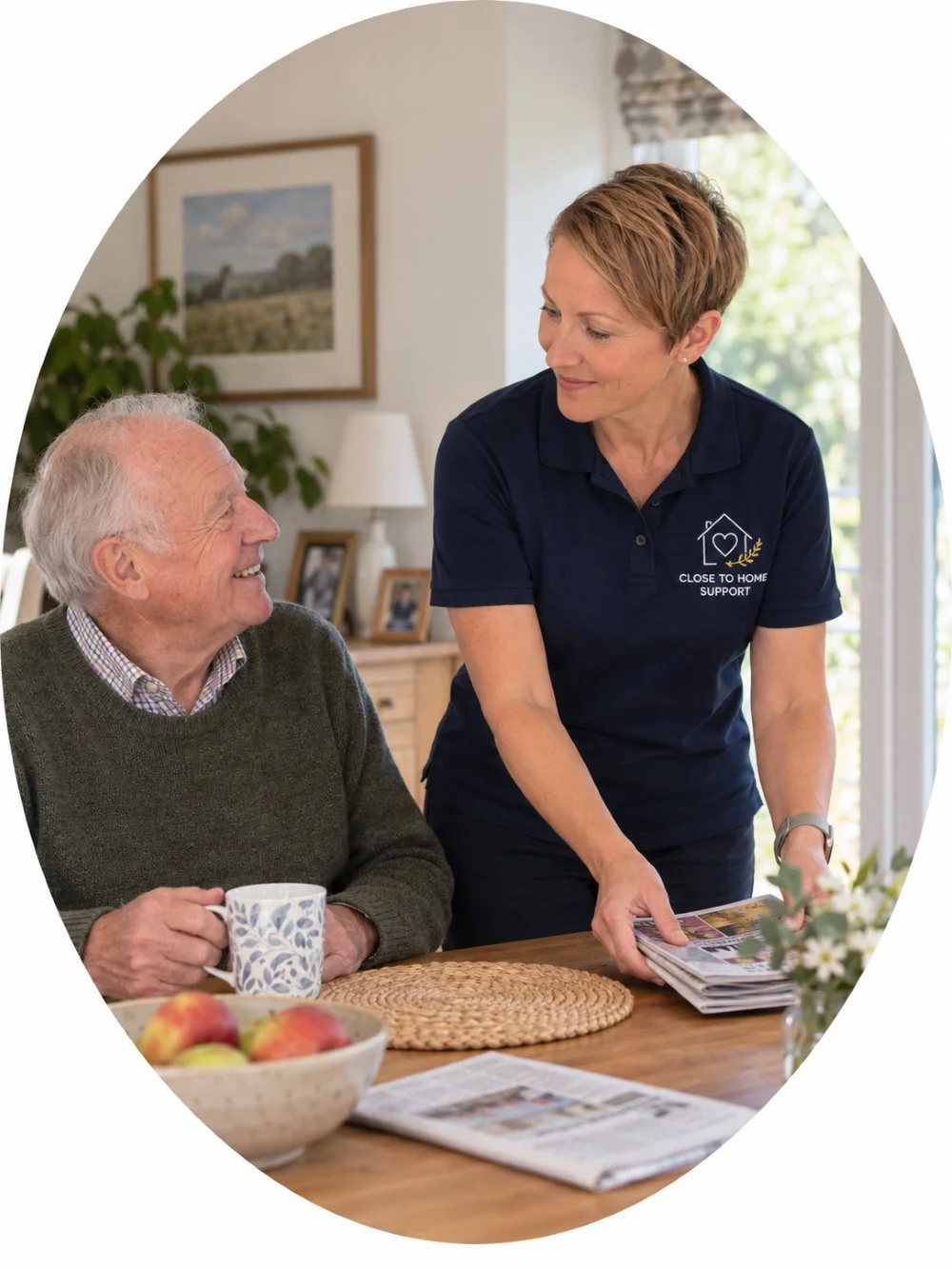 A woman in a navy polo shirt with 'Close to Home Support' logo, assisting an elderly man with reading a magazine at a dining table. The man is smiling, holding a coffee mug, with a bowl of apples nearby.