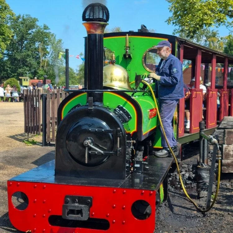 Man dressed in blue work clothes and a cap operating a small, colorful steam train engine at a park or fairground.