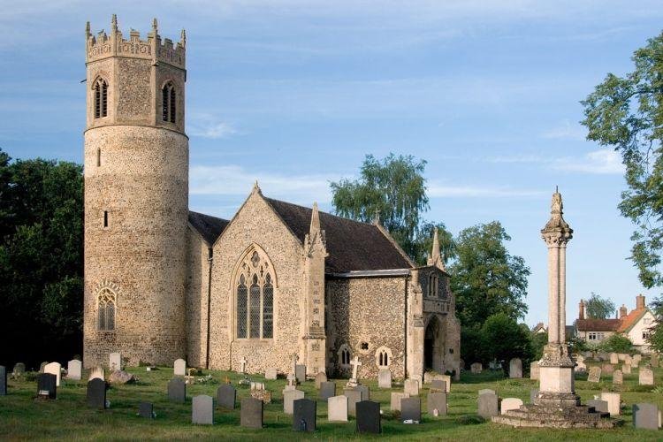 Old stone church with a tower, surrounded by a graveyard with headstones and a monument, set against a blue sky with trees in the background.
