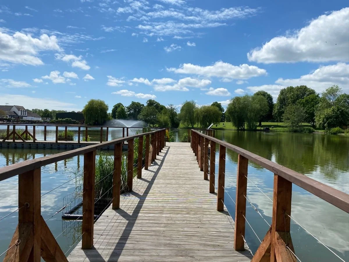 A wooden dock extends over a calm lake on a sunny day with blue sky and scattered clouds. Green trees line the shoreline.