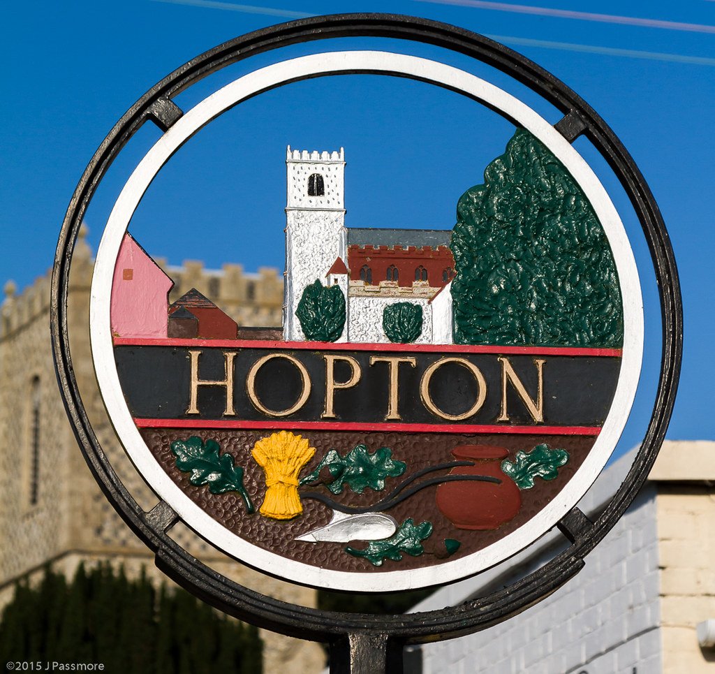 Colorful circular sign for Hopton featuring a church, trees, a yellow wheat sheaf, a pot, and a bird, with background of castle walls.