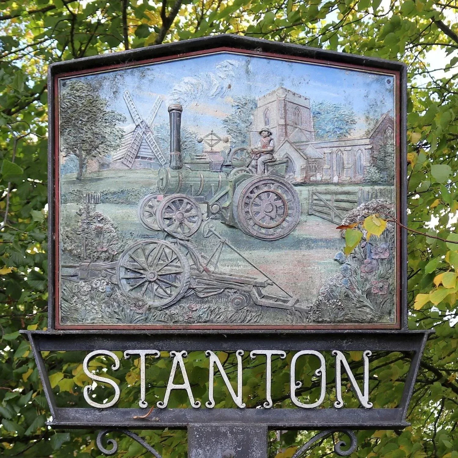 A metal sign with a relief depiction of an old-fashioned train, a church, a windmill, and a farmer riding a tractor. It is surrounded by green trees and has the word 'STANWOOD' at the bottom.