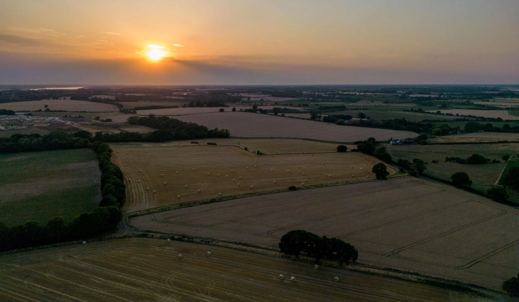 Aerial view of farmland at sunset with fields, trees, and hay bales.