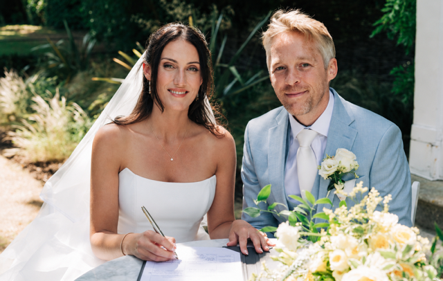 A bride and groom sitting at a table, signing a marriage certificate outdoors during their wedding ceremony. The bride is wearing a white strapless wedding gown and veil, with long dark hair. The groom is in a light blue suit with a white shirt and tie, with blonde hair. There is a bouquet of white and yellow flowers on the table and greenery in the background.