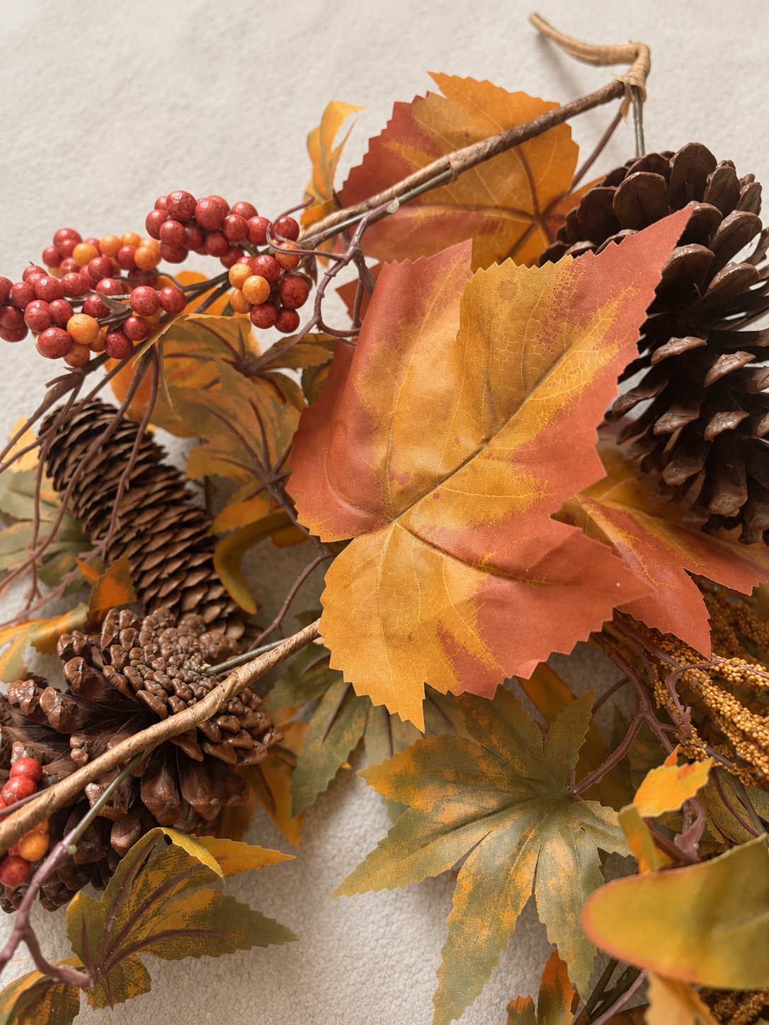Autumn decorative arrangement with orange, yellow, and brown leaves, pinecones, and red berries on a neutral background.