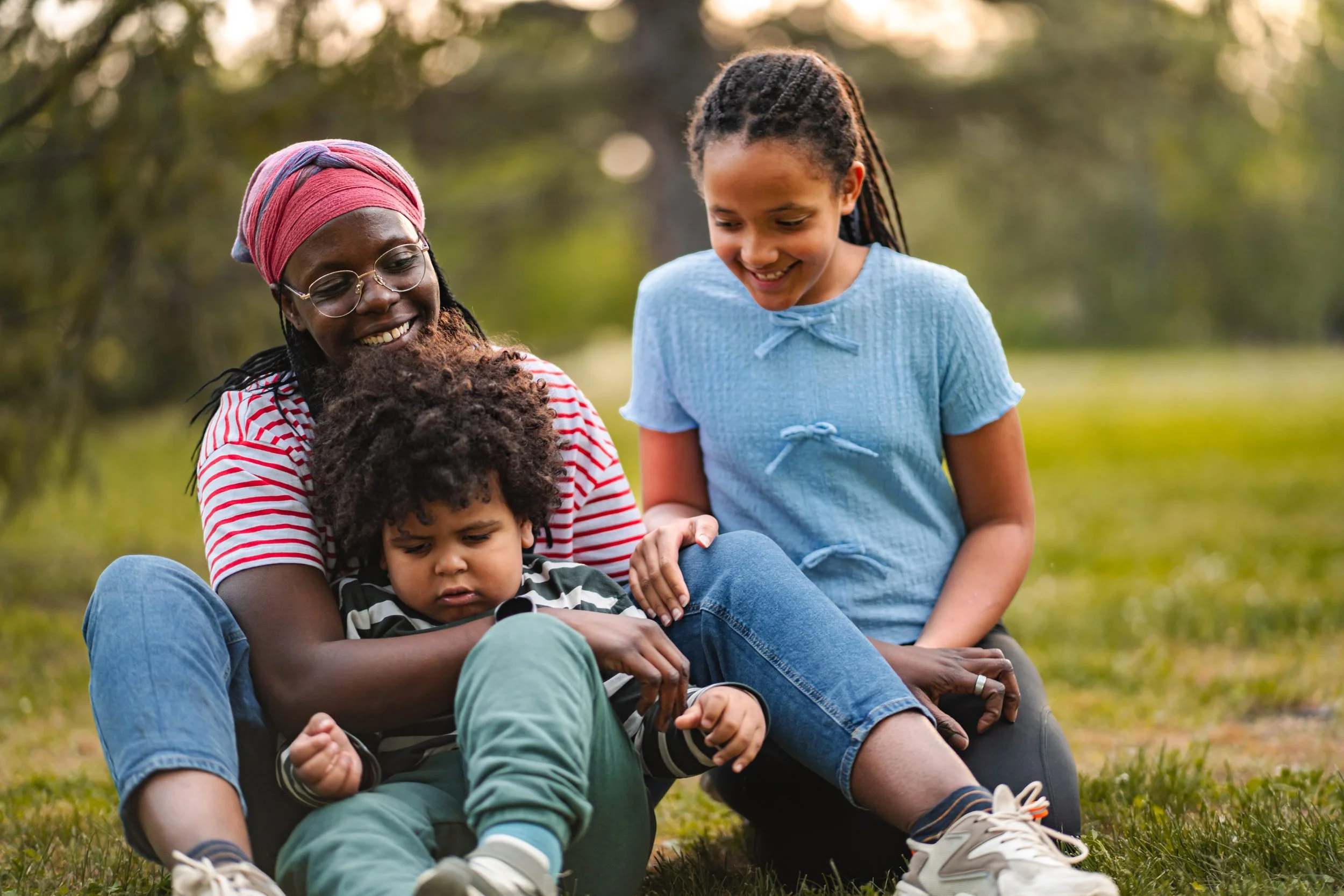 A diverse group of four people sitting outdoors on grass, smiling and spending time together. Two children and two adults, one woman wearing glasses and a pink headscarf, the other woman with braided hair, and children with curly and straight hair. Trees and sunlight in the background.