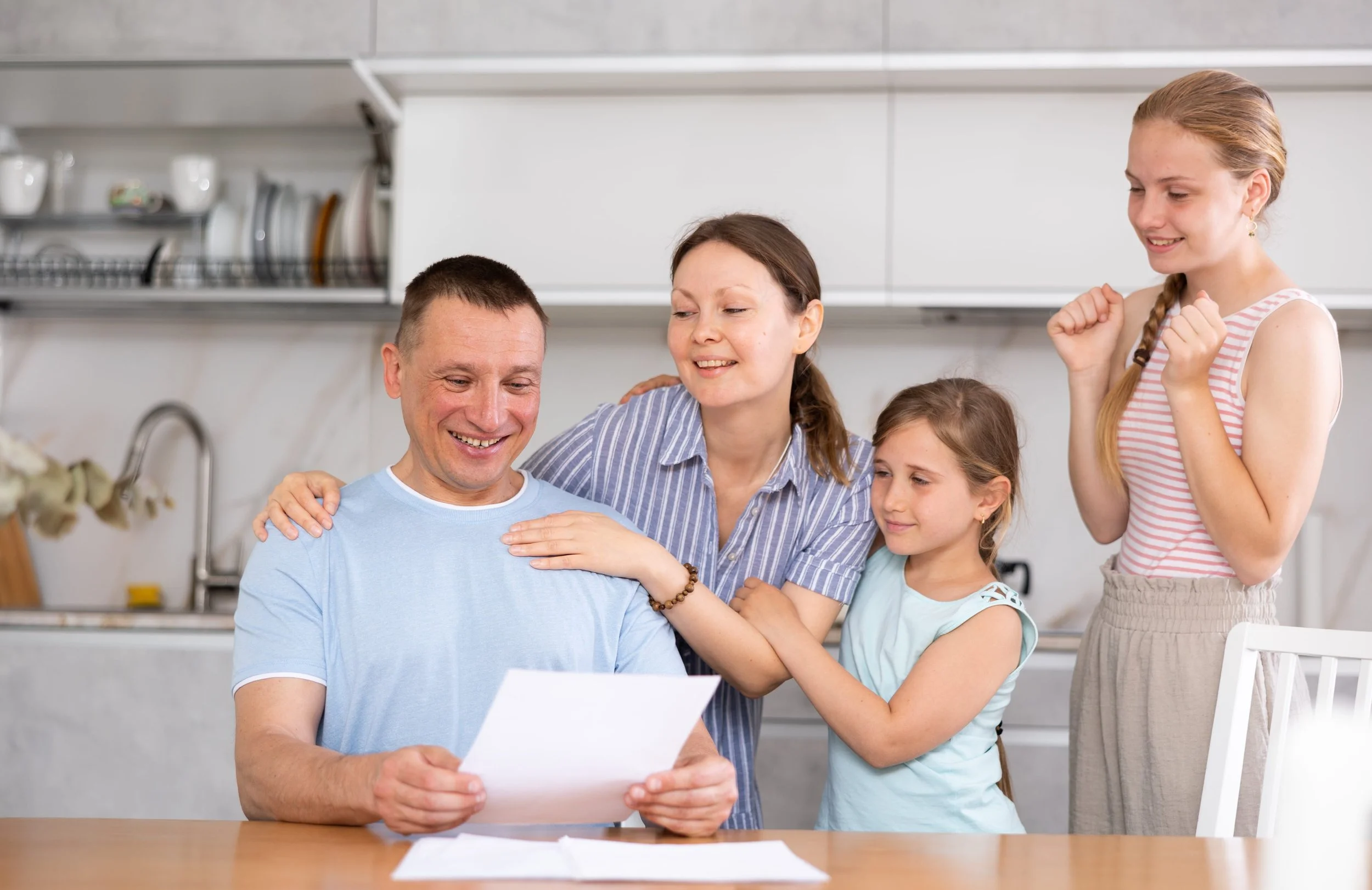 A family gathered around a man sitting at a table, reading a letter, with two children and a woman showing excitement and happiness.