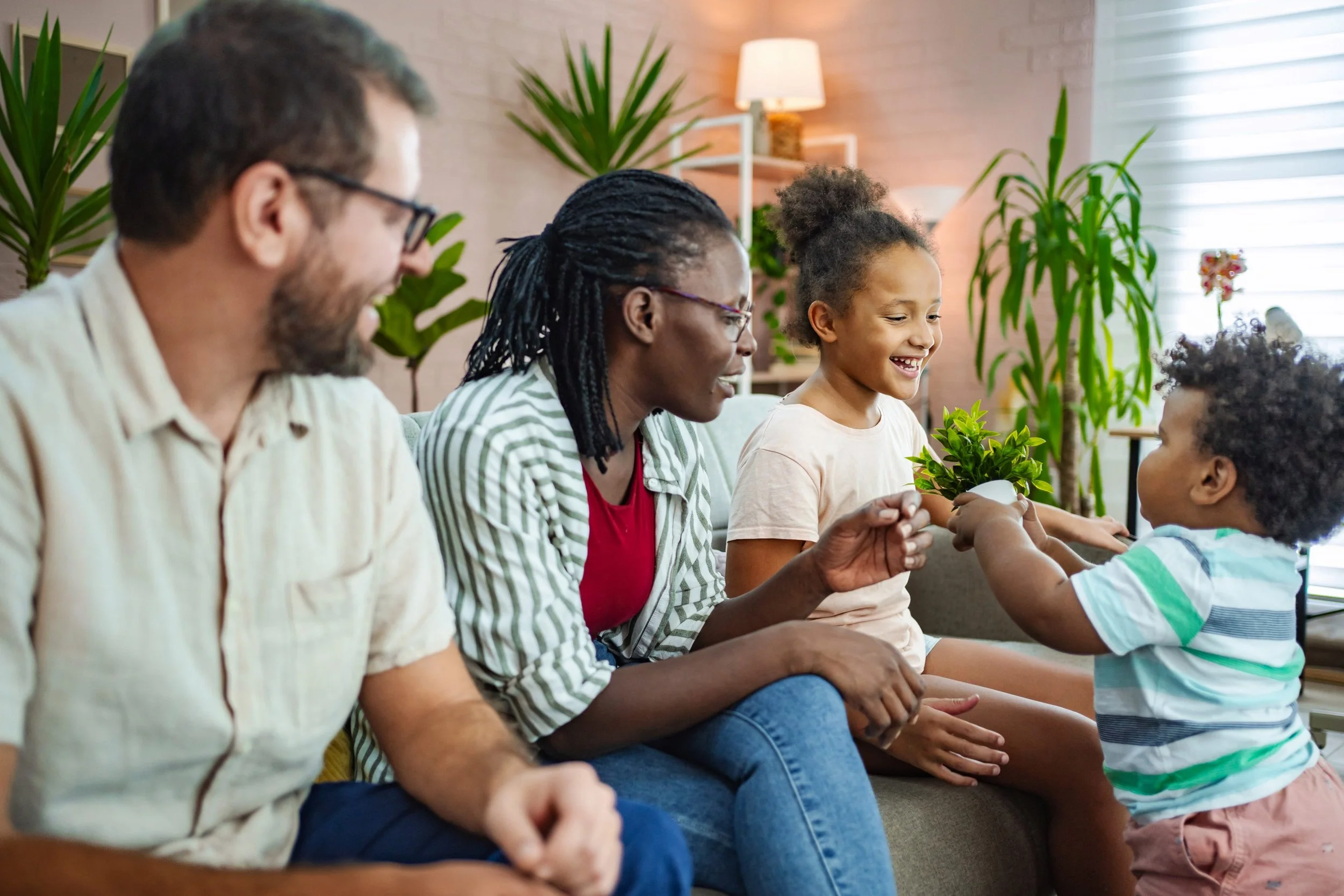 Group of four people sitting on a couch, engaging with two children, surrounded by green plants in a bright room.