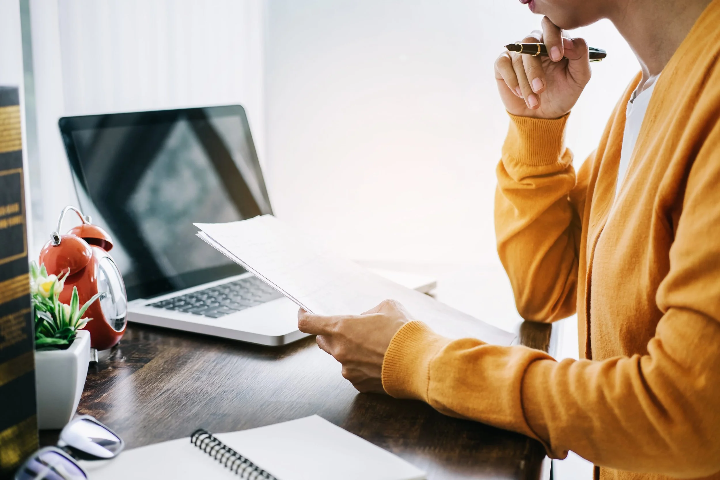 Person in a yellow sweater sitting at a desk, holding a piece of paper and a pen near their face, with a laptop, notebooks, and decorative plants on the desk.
