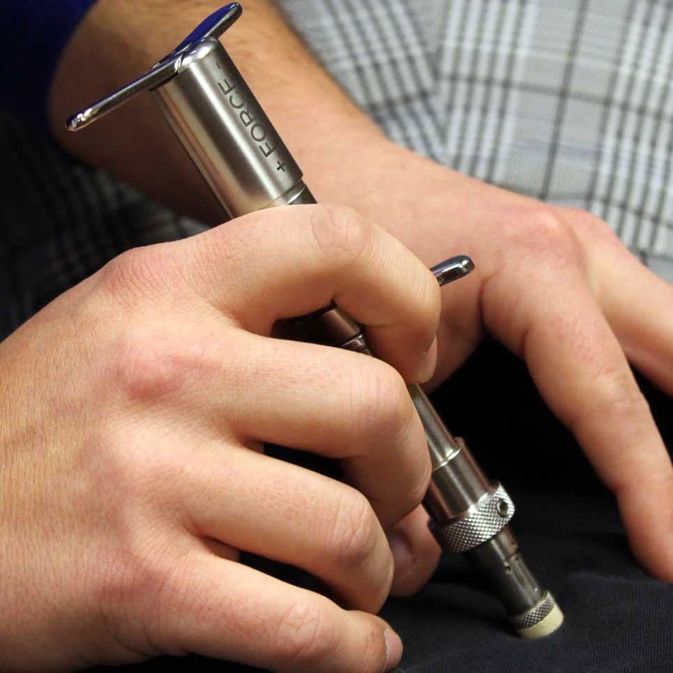 A person's hand holding a metallic tattoo pen over their other hand, with some people wearing plaid clothing visible in the background.