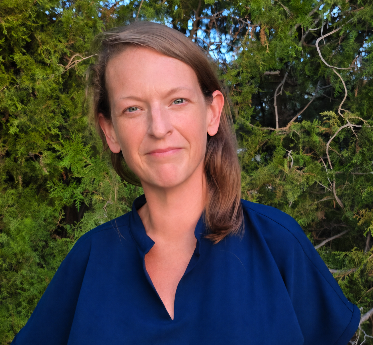 A woman with light skin and light brown hair wearing a blue top, standing outdoors in front of green foliage, smiling softly at the camera.