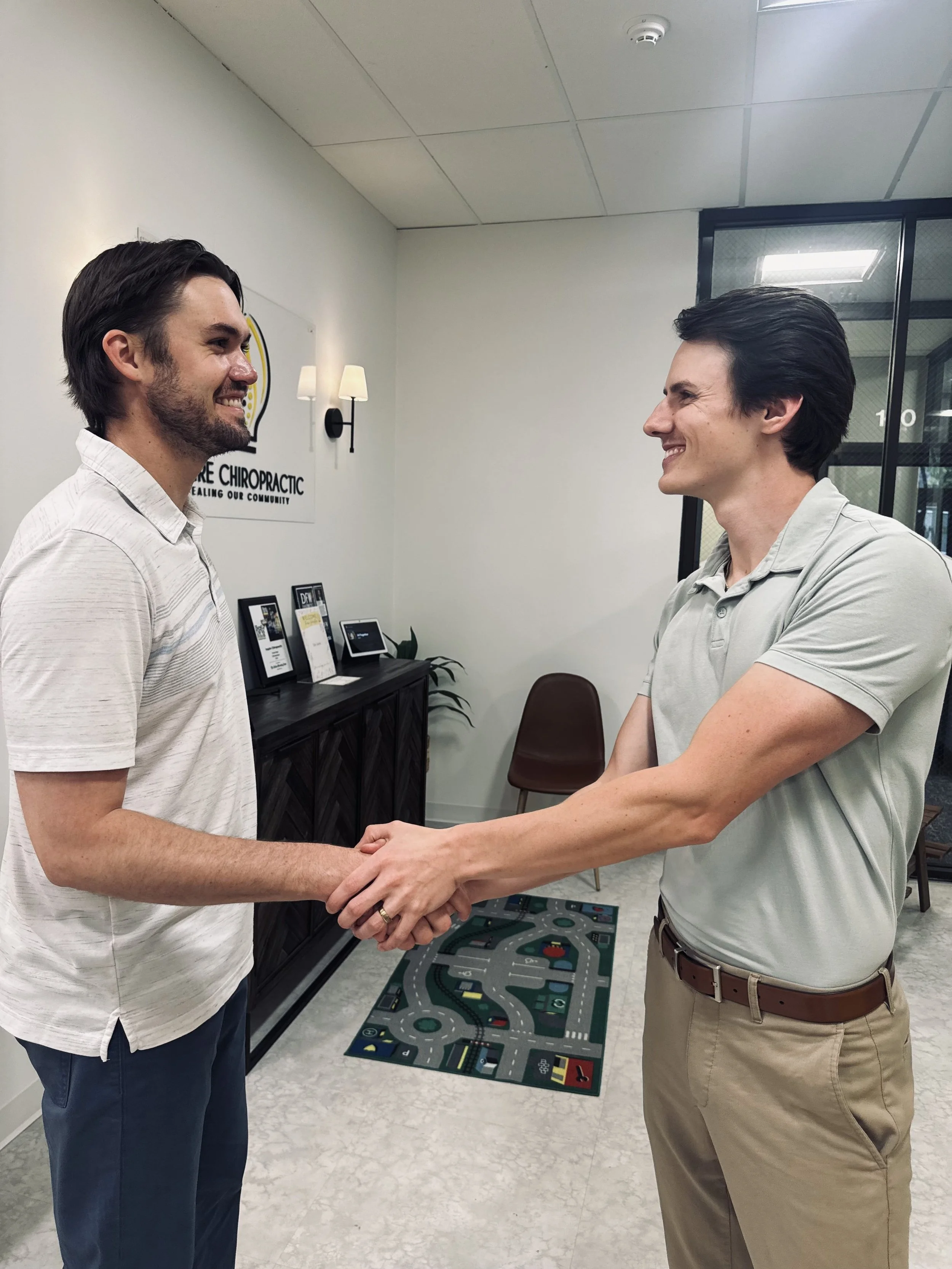 Two men are smiling and shaking hands in an office. One man has dark hair and is wearing a light striped polo shirt. The other man has dark hair and is wearing a light color polo shirt with khaki pants. There are framed certificates or awards on a table behind them and a children's play mat on the floor.