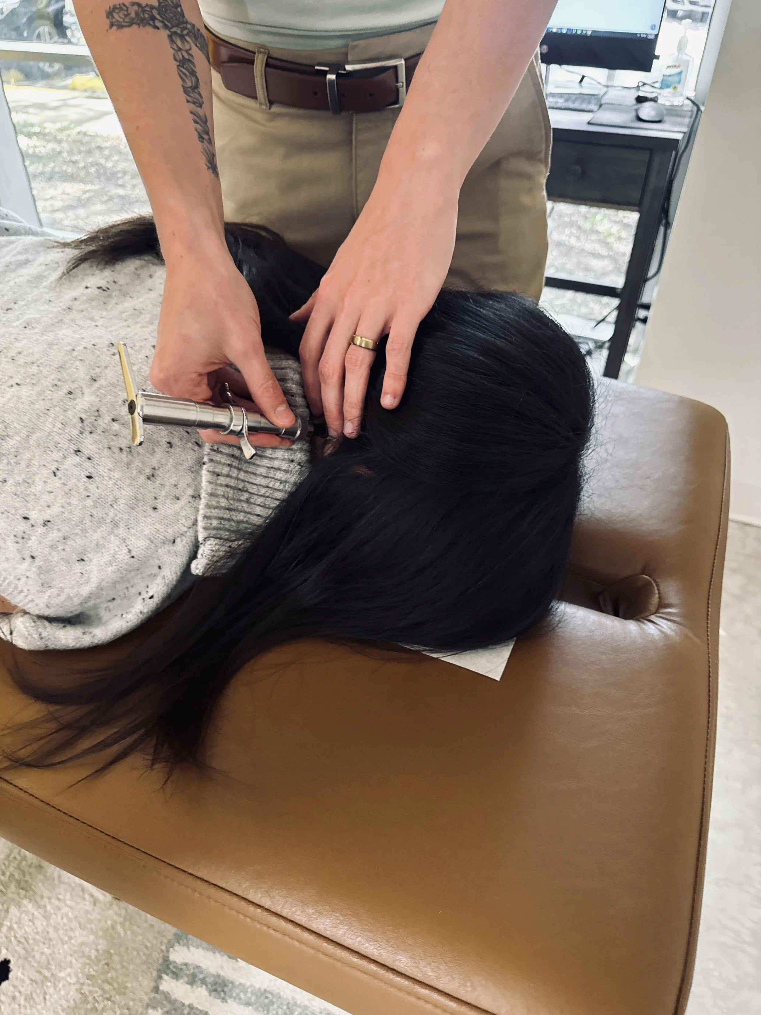 A person receiving a haircut at a salon, lying face down on a cushioned chair with long dark hair, while the hairdresser trims the hair using scissors.