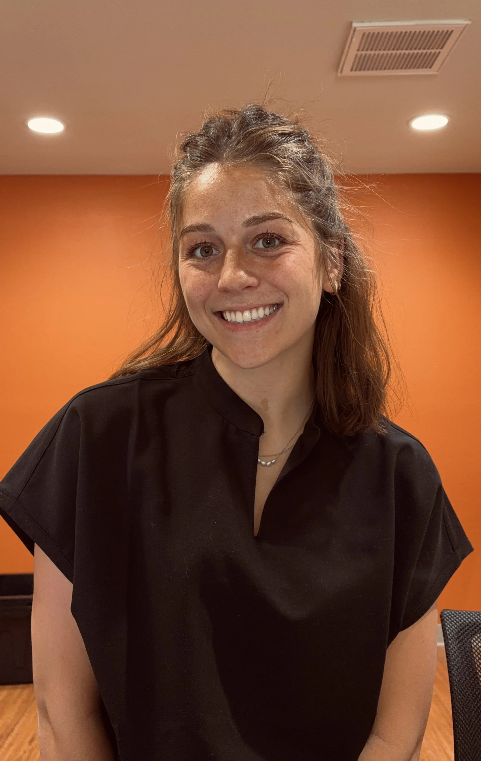 A smiling woman with light brown hair, tied back, wearing a black shirt and a necklace, standing in front of an orange wall with ceiling lights.