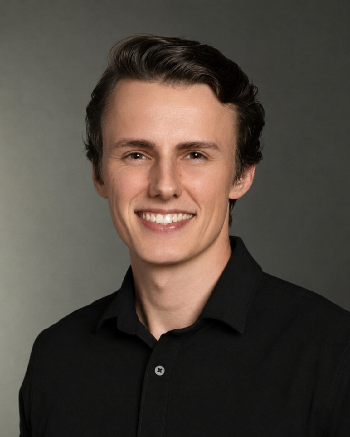A young man with short, wavy brown hair, wearing a black collared shirt, smiling against a plain gray background.
