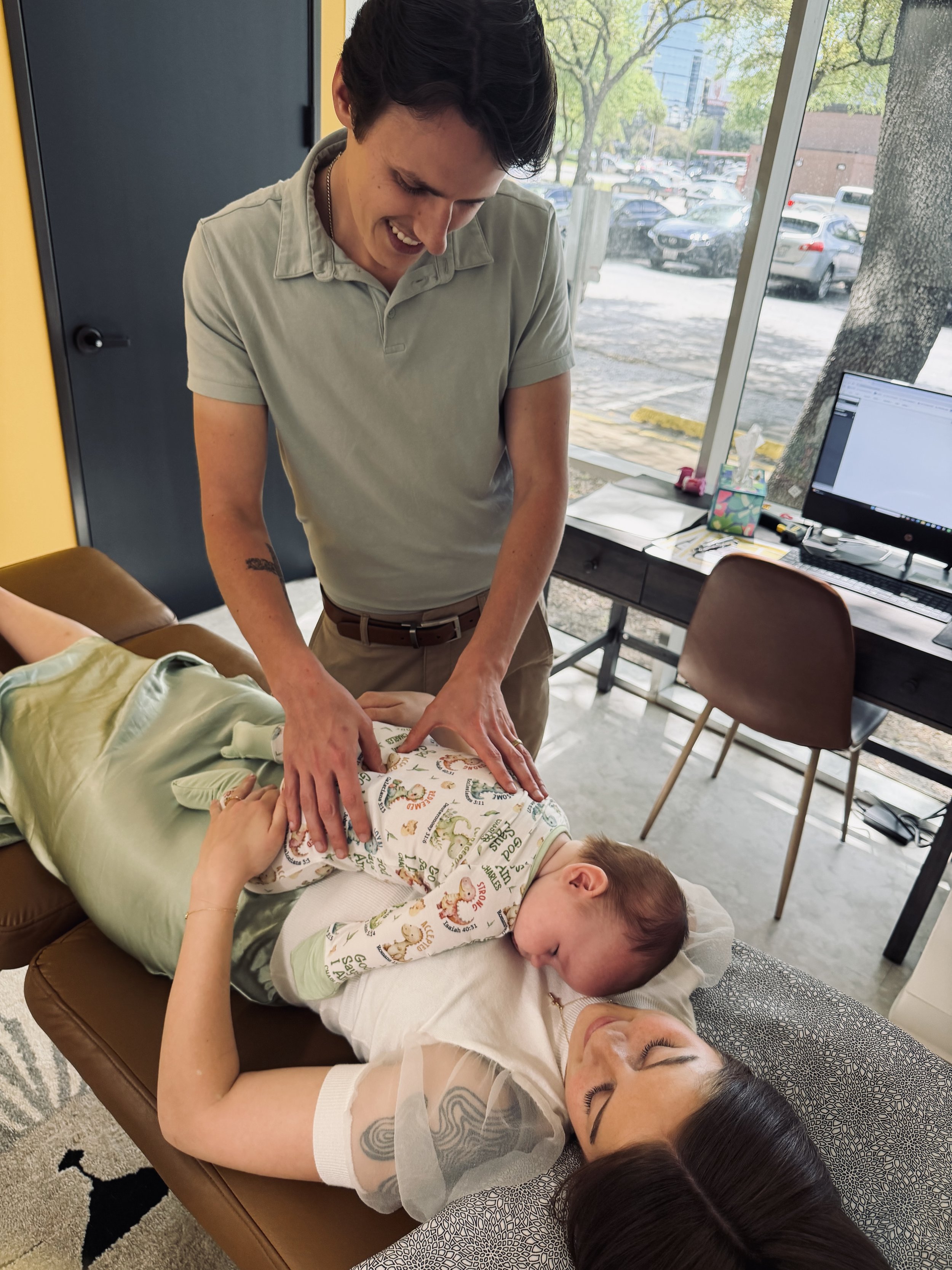A woman giving a massage to a young boy who is lying face down on a massage table, with another woman and a young girl lying on the floor nearby, in a room with large windows and parked cars outside.