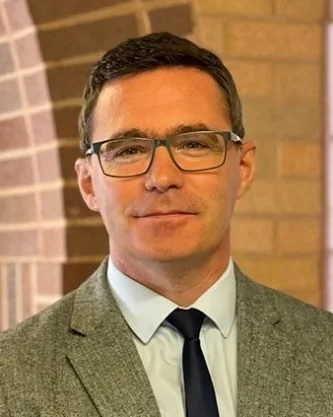 A man wearing glasses, a white shirt, a black tie, and a gray suit jacket standing in front of a brick wall.