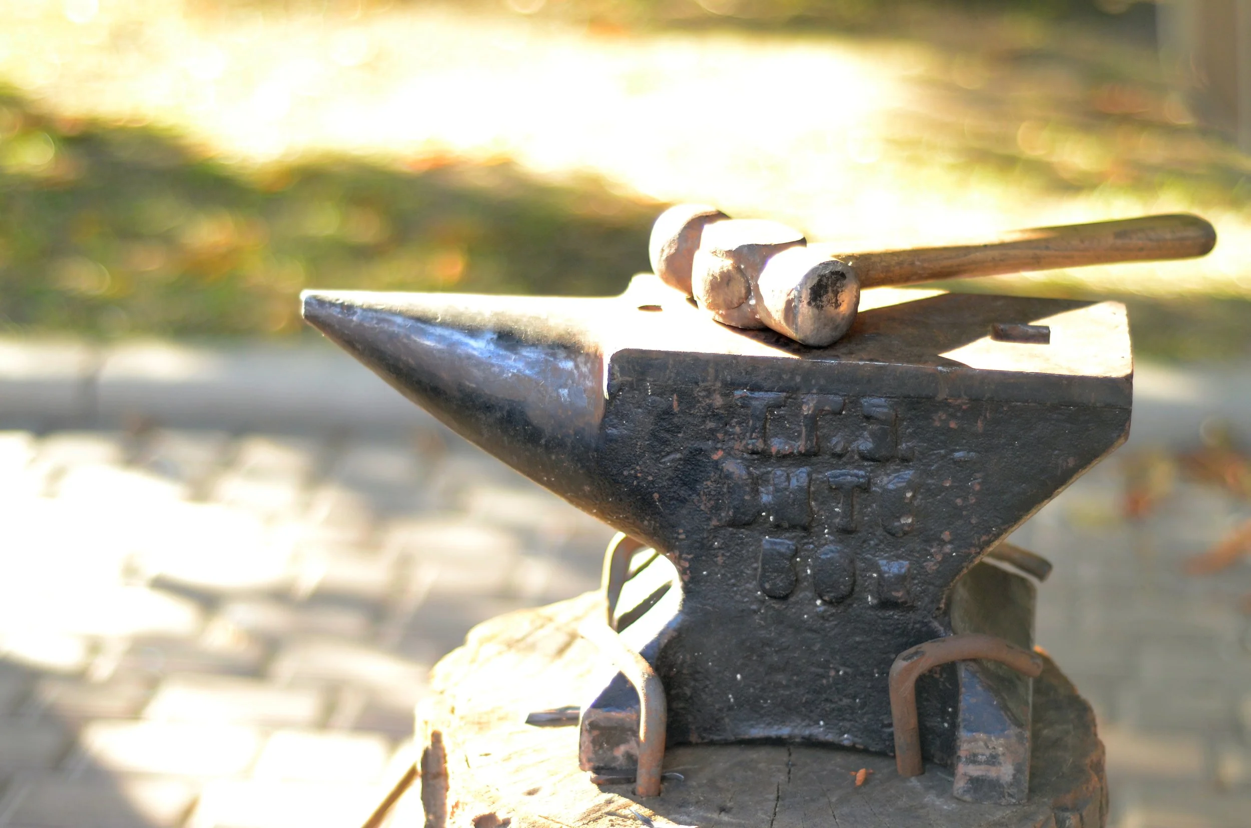 Close-up of a blacksmith's anvil with two metal hammers resting on it, outdoors with blurred greenery in the background.