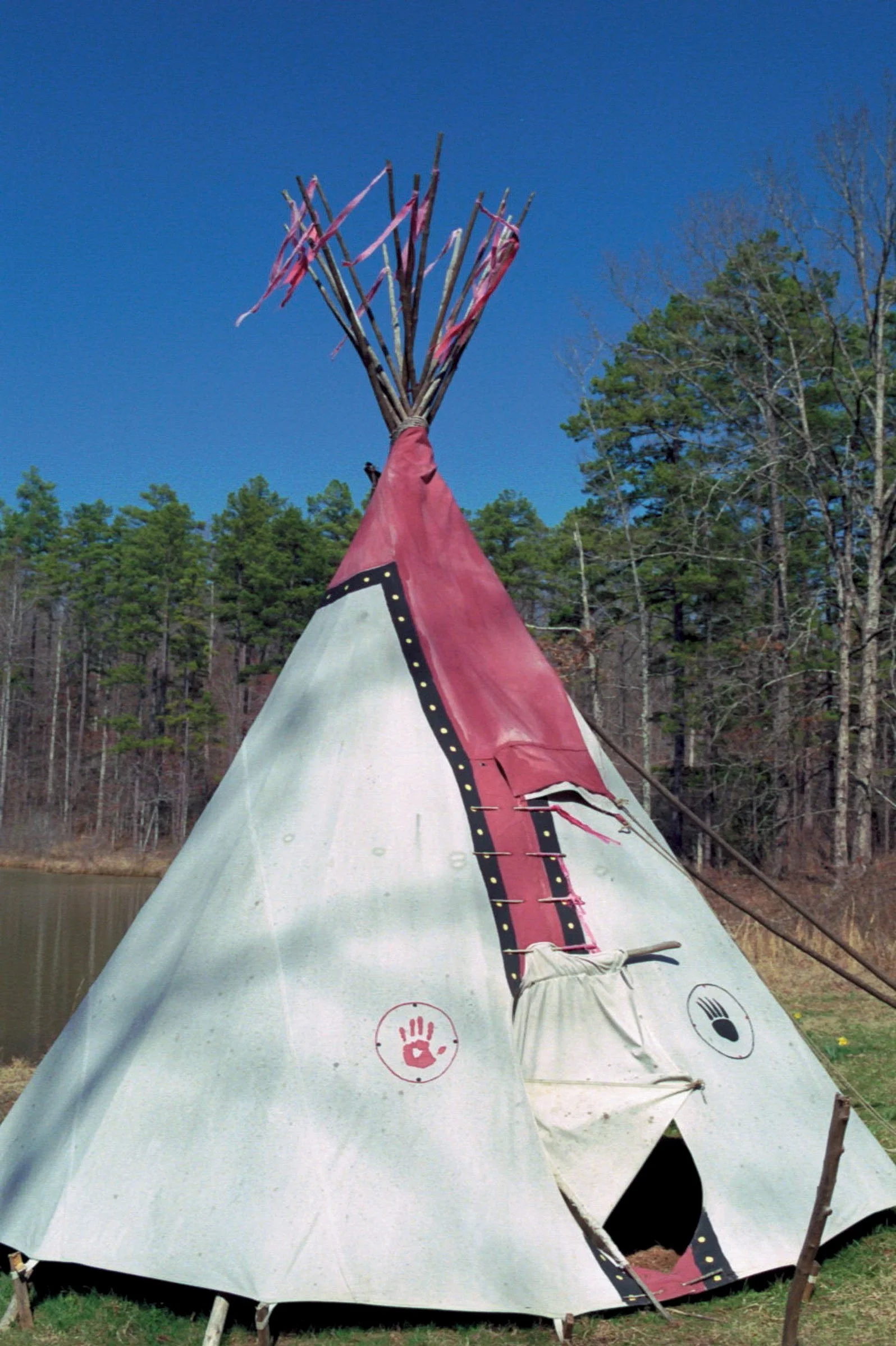 A white teepee tent with pink and black accents, set up outdoors near a body of water with trees and a blue sky in the background.