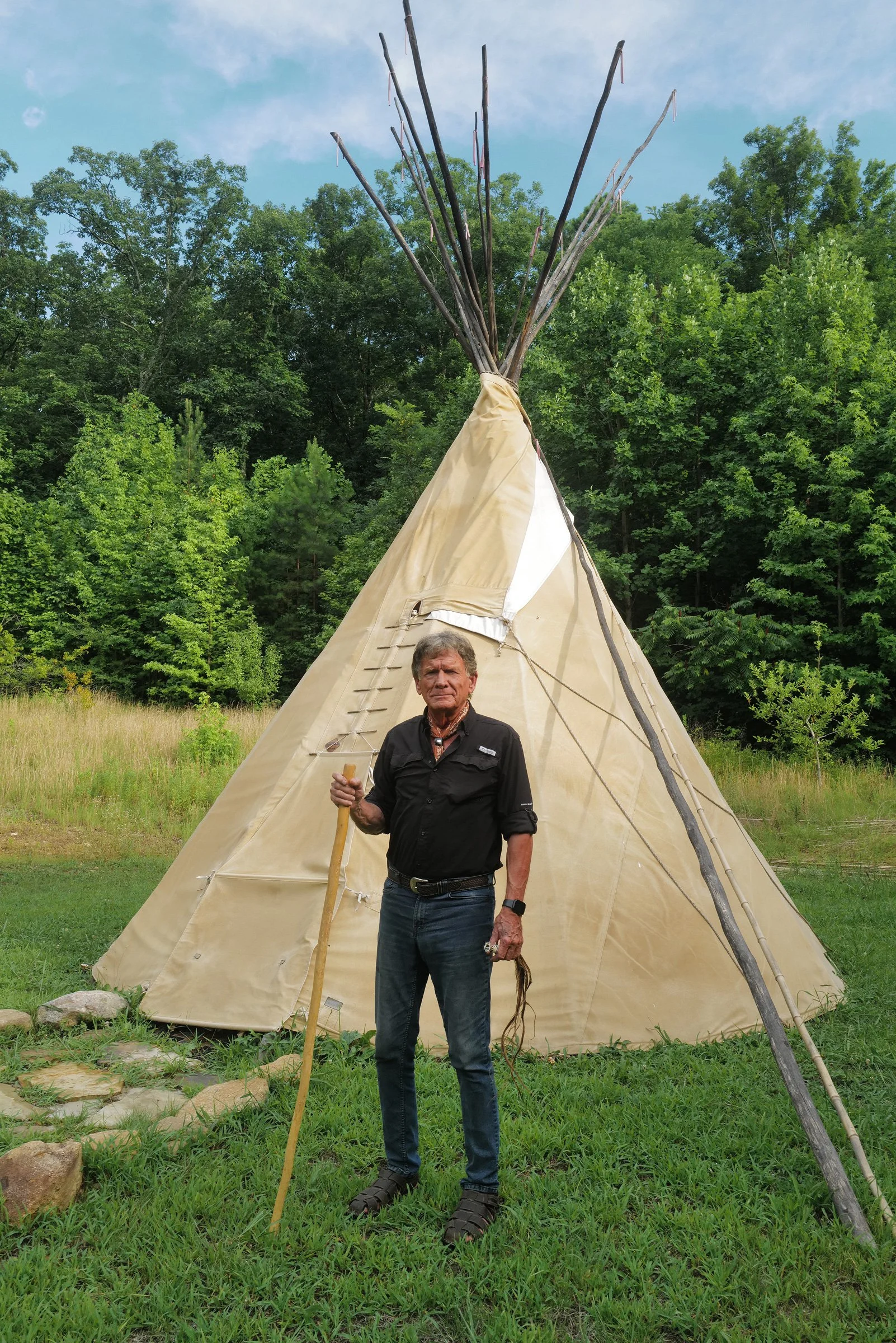 A man standing outside a large beige tipi tent holding a long stick, with a forest background and a partly cloudy sky.