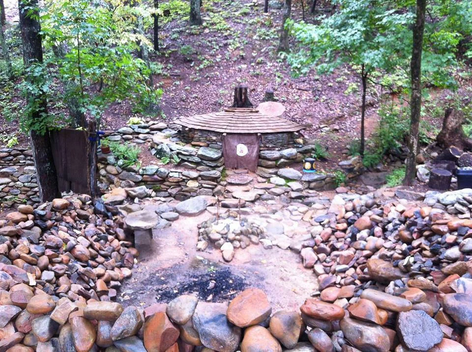 A small, rustic outdoor scene in a wooded area with a stone and wood structure resembling a tiny house, surrounded by rocks and trees.